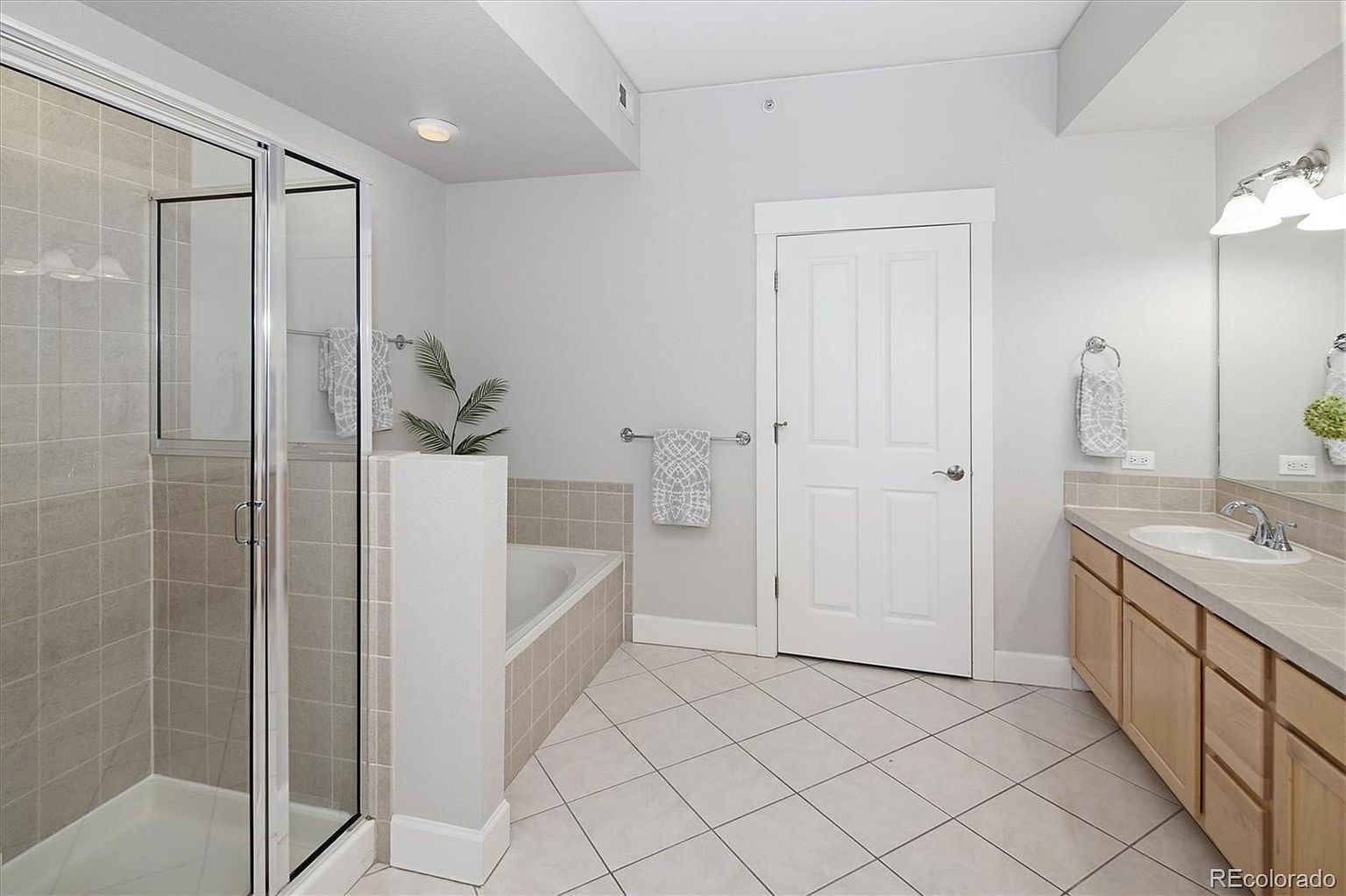This spacious primary bathroom features a clean, neutral aesthetic with light-colored tile flooring and walls. The room includes a glass-enclosed walk-in shower, a separate soaking tub, and a long vanity with light wood cabinetry and a large mirror. The layout is functional and bright, illuminated by overhead lighting and a vanity light fixture.