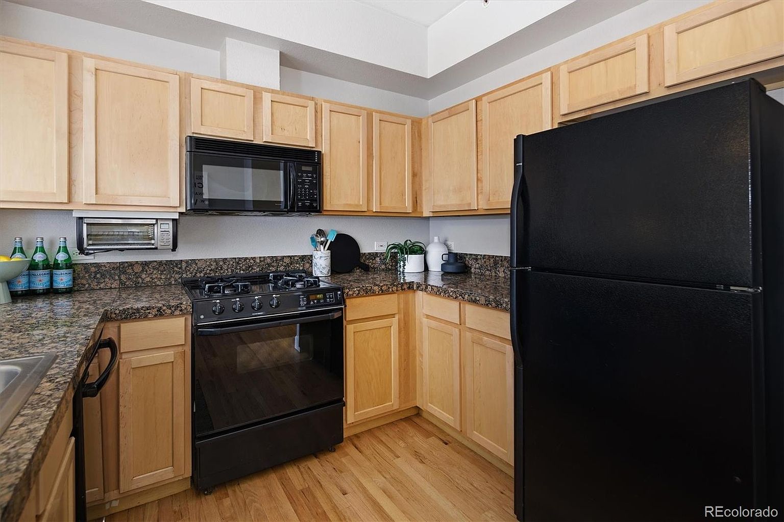 This kitchen features light wood cabinetry paired with dark granite countertops and a black appliance suite, including a gas range and a microwave. The space is efficiently laid out in an L-shape, providing a functional workspace with a neutral color palette. The perspective is a straight-on eye-level shot that captures the warmth of the wood tones and the contrast of the dark fixtures.