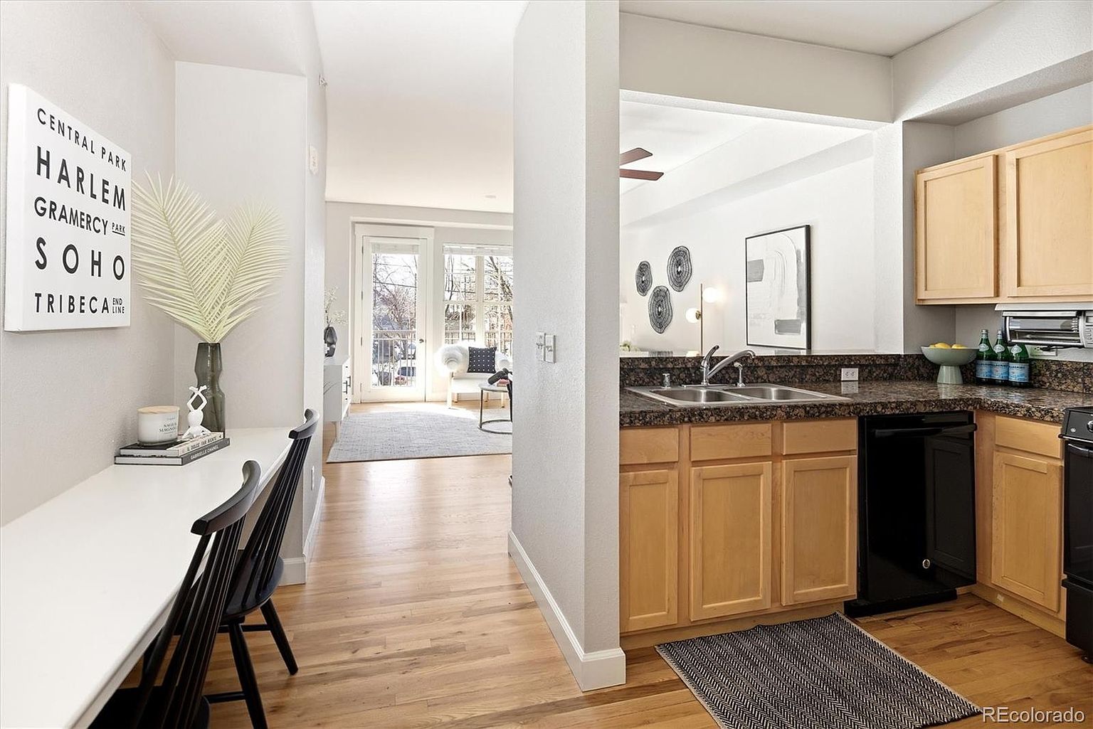 This view captures a functional kitchen area transitioning into a bright living space, featuring light wood cabinetry, dark granite countertops, and a black dishwasher. To the left, a sleek built-in breakfast bar with black chairs provides a cozy dining nook, while the open floor plan leads the eye toward a sunlit patio door. The overall aesthetic is clean and modern, emphasizing a seamless flow between the culinary workspace and the living area.