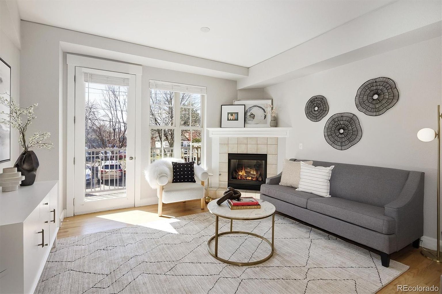 This bright and inviting living room features a contemporary gray sofa, a marble-topped coffee table, and a cozy armchair accented with a faux-fur throw. A fireplace serves as the focal point, complemented by minimalist wall art and a neutral-toned area rug that ties the space together. The room is bathed in natural light from the adjacent glass door leading to a balcony, creating an airy and sophisticated atmosphere.