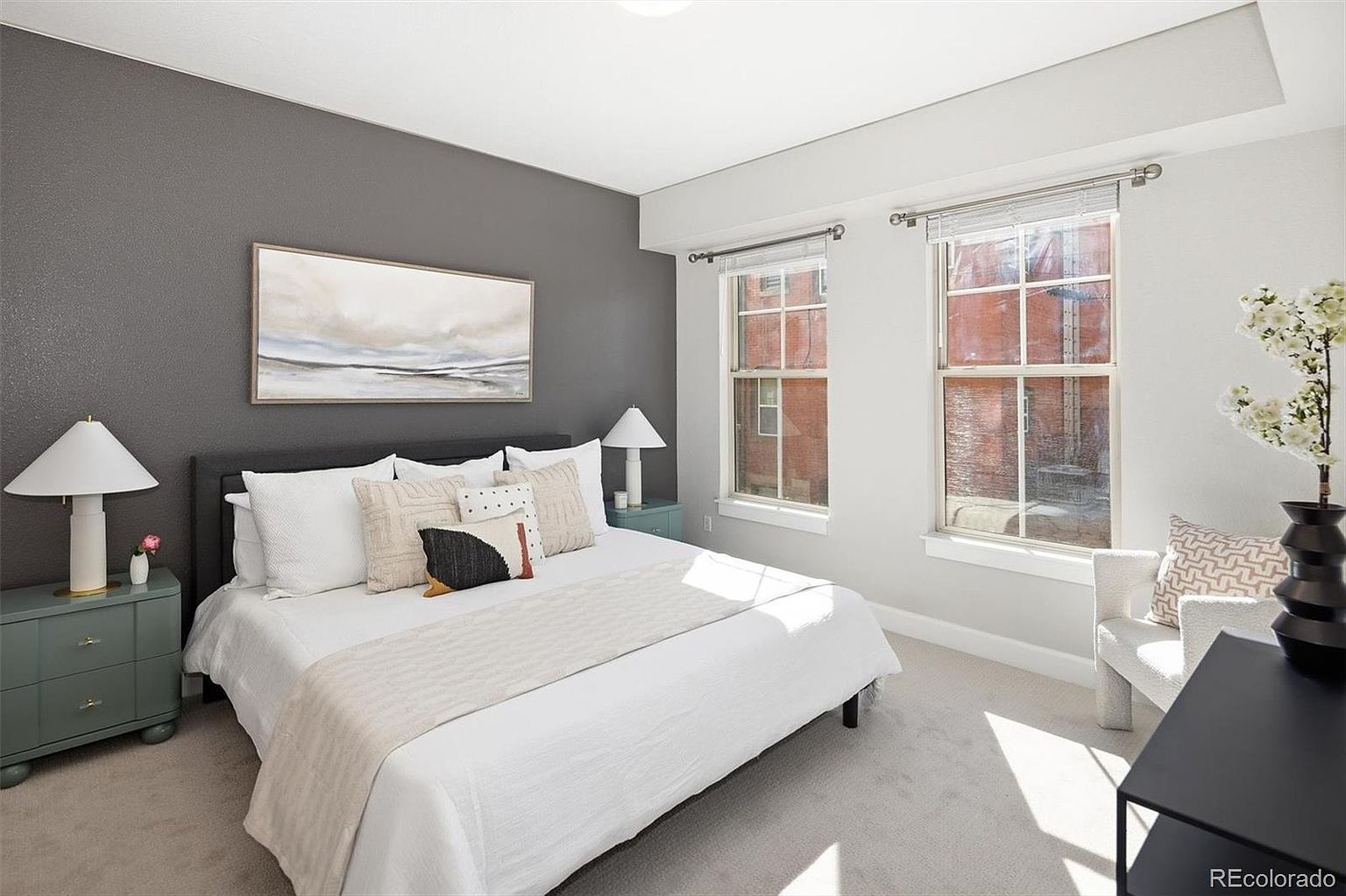 This bright and inviting primary bedroom features a modern aesthetic with a striking dark gray accent wall behind the bed. The room is furnished with a plush king-sized bed, two matching sage-green nightstands with elegant lamps, and a cozy armchair near the window. Natural light streams through the two large windows, creating a serene and airy atmosphere perfect for relaxation.