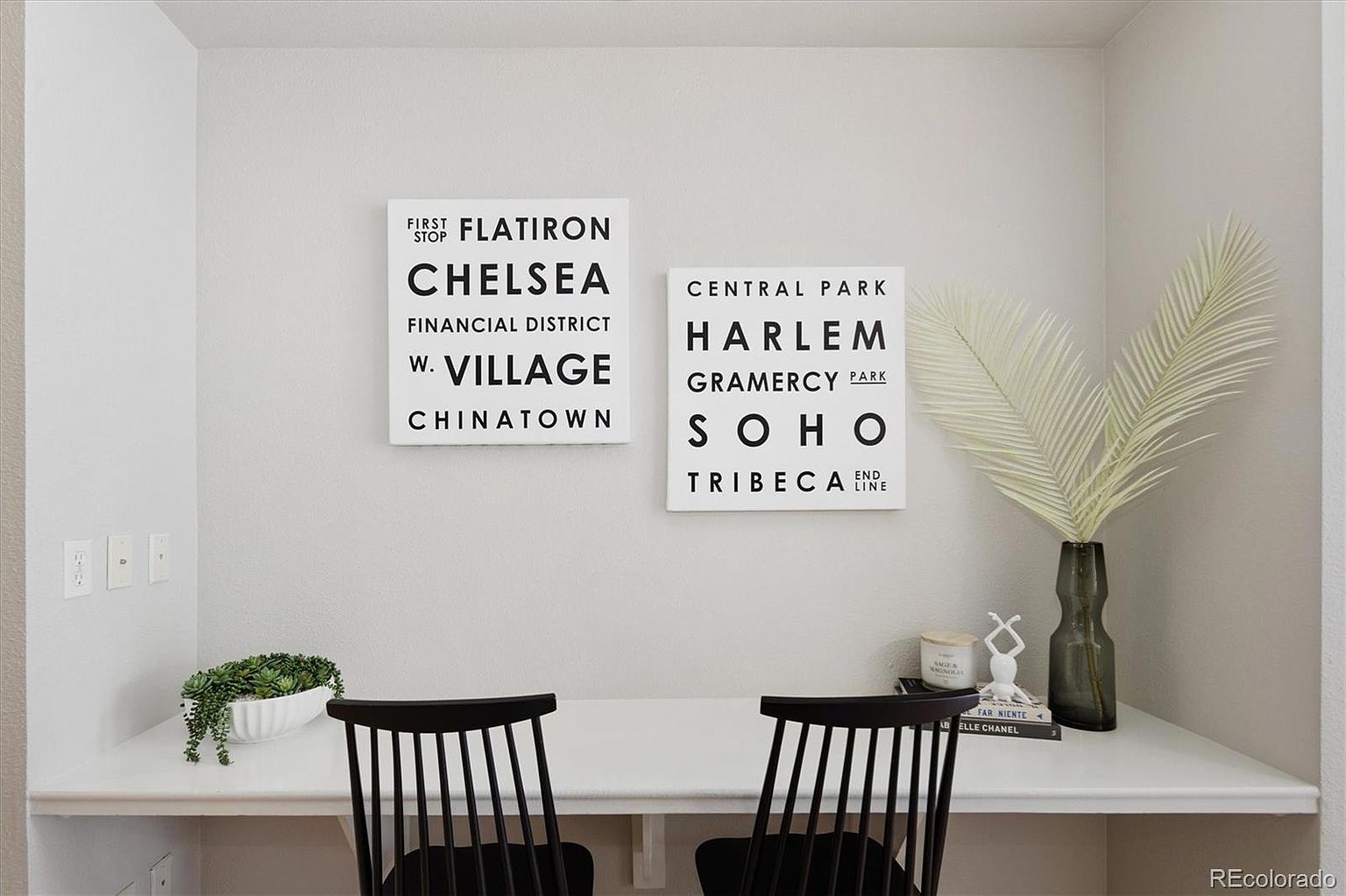 This cozy home office nook features a clean, minimalist desk setup with two black chairs and a pair of modern, subway-style wall art pieces depicting New York City neighborhoods. The space is accented by a small potted succulent, a decorative white sculpture, and a tall glass vase with a palm leaf, creating a bright and inviting workspace. The neutral color palette and simple decor provide a contemporary and organized aesthetic perfect for a productive home environment.