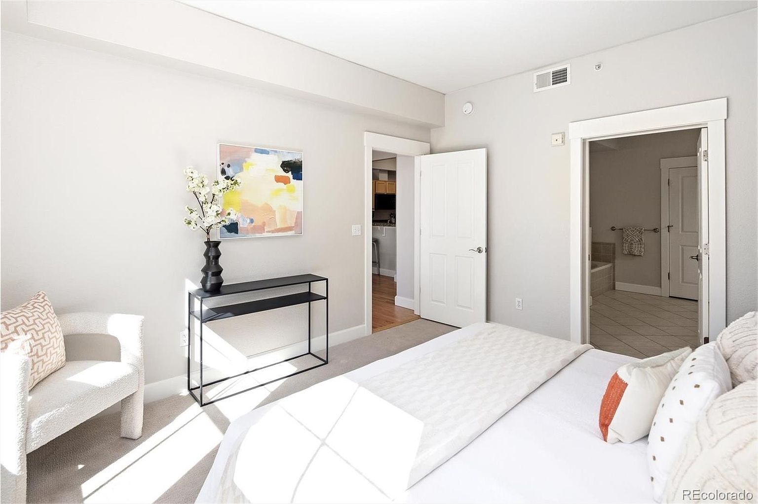 This bright and airy primary bedroom features neutral tones, plush carpeting, and a clean, modern aesthetic. The room includes a comfortable bed with textured pillows, a sleek black console table topped with a floral arrangement, and a piece of abstract wall art. A doorway leads into an adjoining bathroom, while another open doorway provides a glimpse into a secondary living space, creating an open and inviting flow.