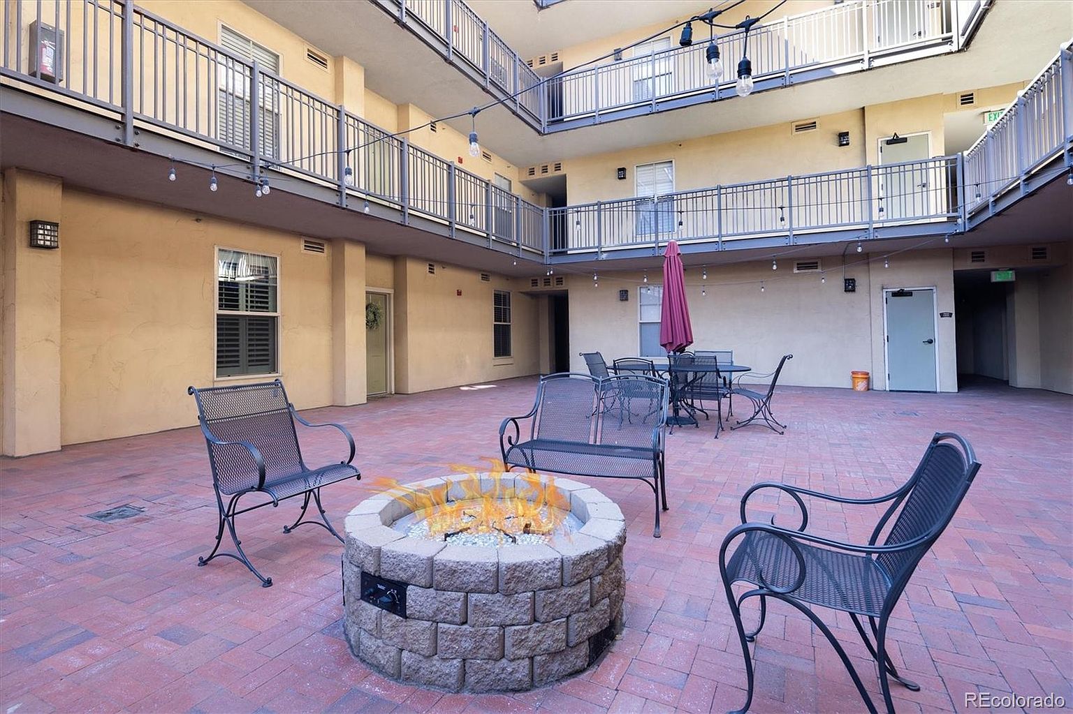 This image captures a communal courtyard area within an apartment complex, featuring a central stone fire pit surrounded by metal patio furniture. The space is enclosed by multi-story building wings with balconies, creating a private, urban oasis atmosphere. The brick-paved ground and string lighting overhead contribute to a cozy, inviting environment perfect for social gatherings.