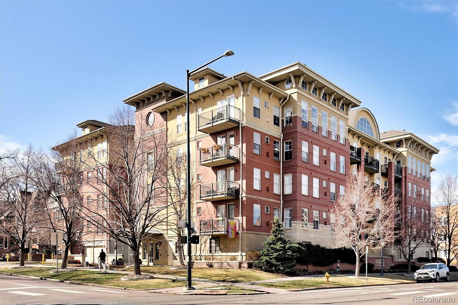 This image captures a multi-story brick and stucco apartment building from a street-level perspective, showcasing its classic architectural design with multiple balconies. The structure features a prominent corner profile, large windows, and a decorative roofline, set against a clear blue sky. A mature tree in the foreground and a nearby street lamp add depth to the urban residential setting.