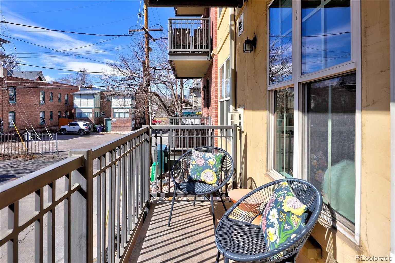 This image captures a cozy, elevated balcony space featuring two modern, dark-framed woven chairs adorned with vibrant floral cushions. The balcony is enclosed by a simple metal railing and overlooks a paved parking area and neighboring brick buildings, offering an urban residential perspective. The scene is bathed in bright, natural daylight, creating an inviting and relaxed outdoor nook.