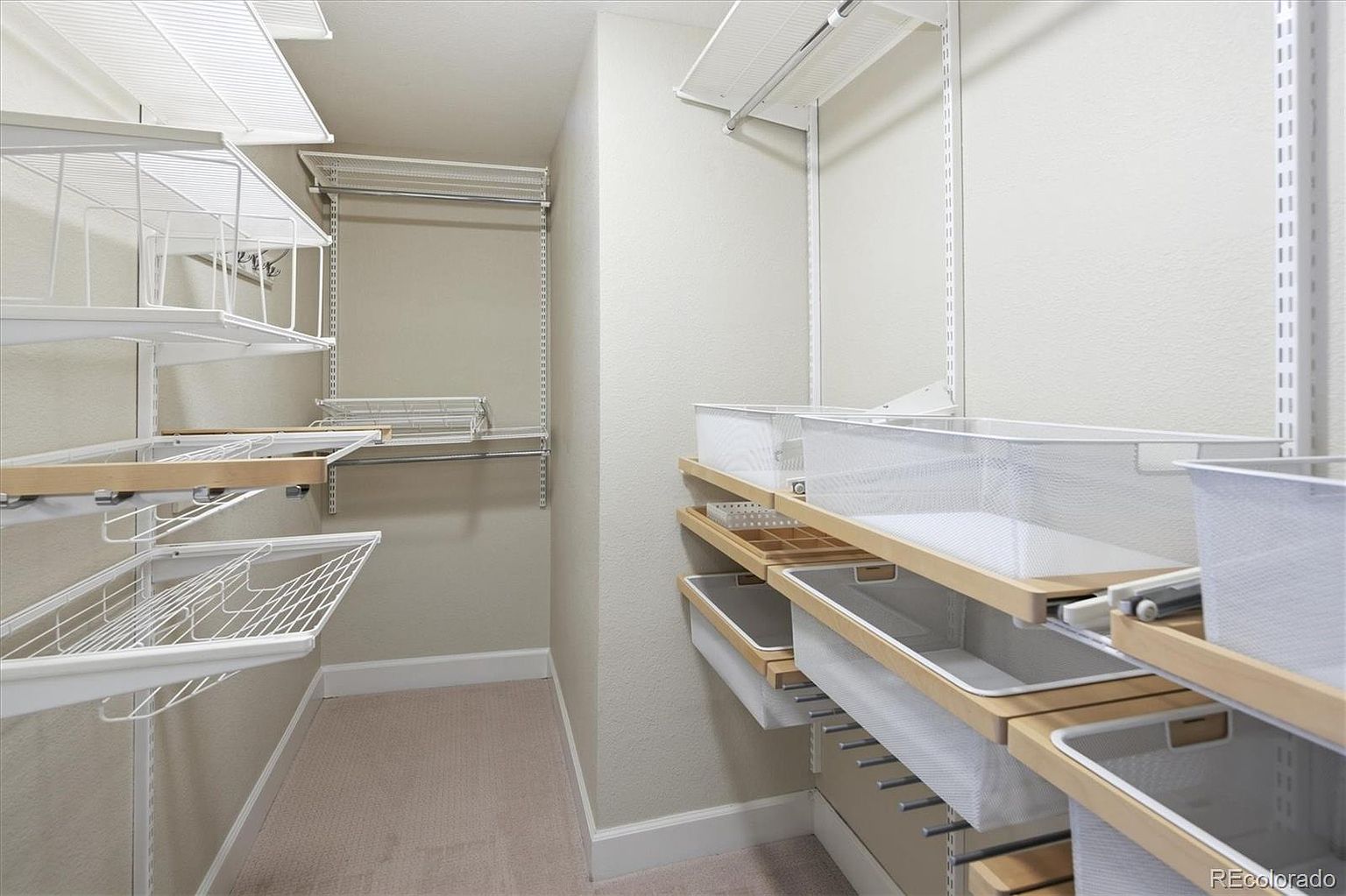 This organized walk-in closet features a modular white wire shelving system with integrated wooden trim accents. The space includes multiple hanging rods, open shelving, and pull-out wire baskets, providing ample storage and a clean, functional aesthetic. The neutral wall color and carpeted flooring create a bright and inviting atmosphere.