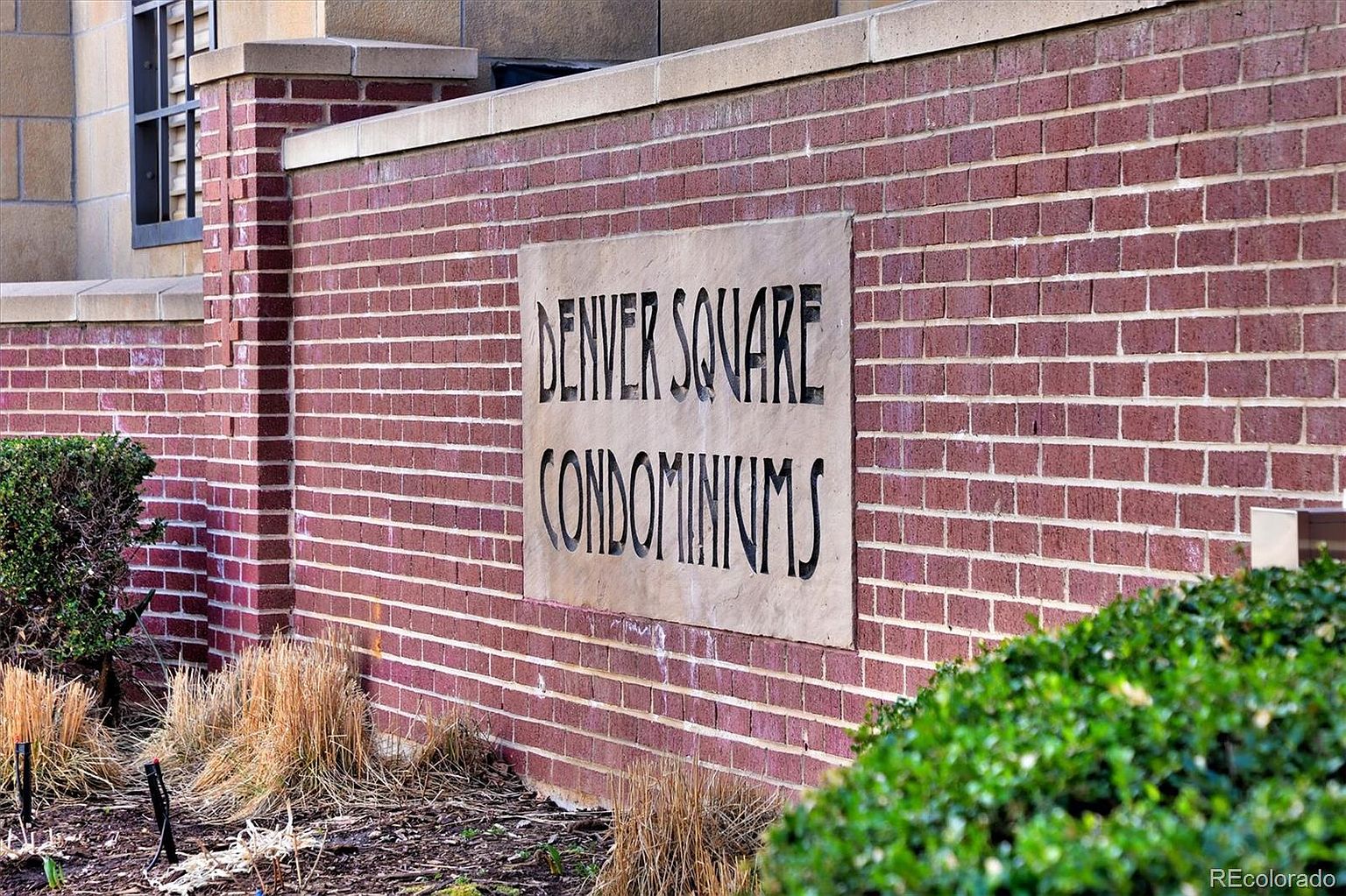 This image features a close-up, angled shot of a brick wall displaying a stone plaque that reads 'DENVER SQUARE CONDOMINIUMS' in bold, dark lettering. The scene includes a manicured green bush in the foreground and dry, ornamental grasses, suggesting a well-maintained community entrance. The perspective is low and focused, highlighting the textured masonry and professional signage of the property.