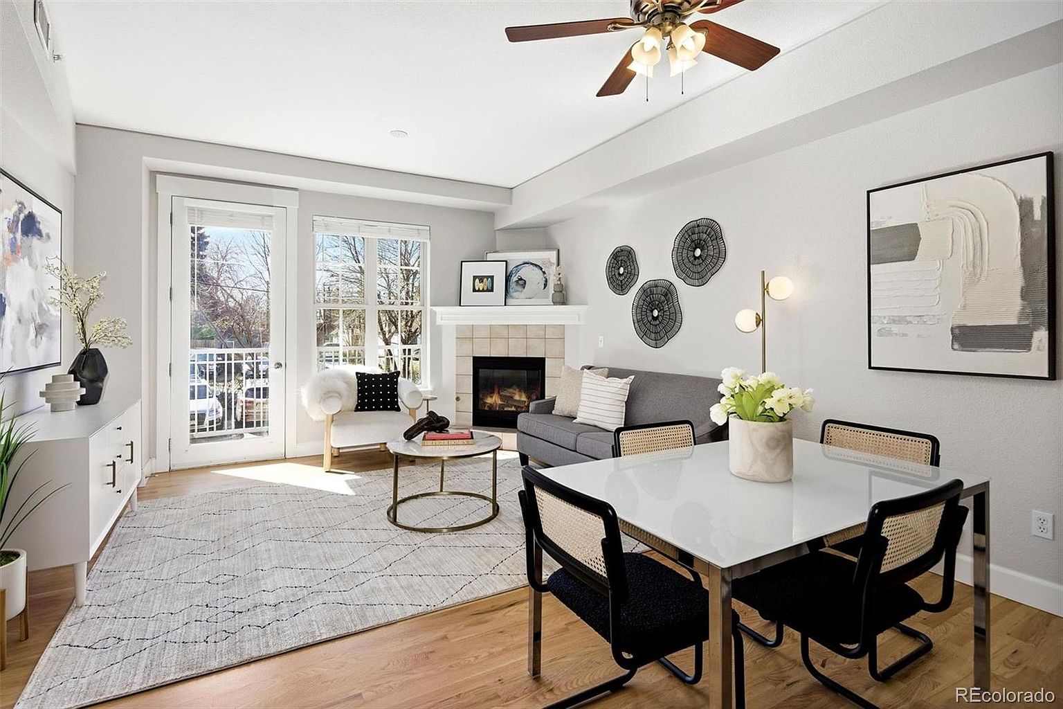 This bright and inviting open-concept living and dining area features a modern aesthetic with light hardwood floors and neutral walls. The space includes a cozy seating area with a grey sofa and fireplace, alongside a contemporary dining set with cane-back chairs, all illuminated by natural light from the adjacent balcony door. The room is tastefully decorated with abstract wall art and a textured area rug, creating a cohesive and stylish living environment.