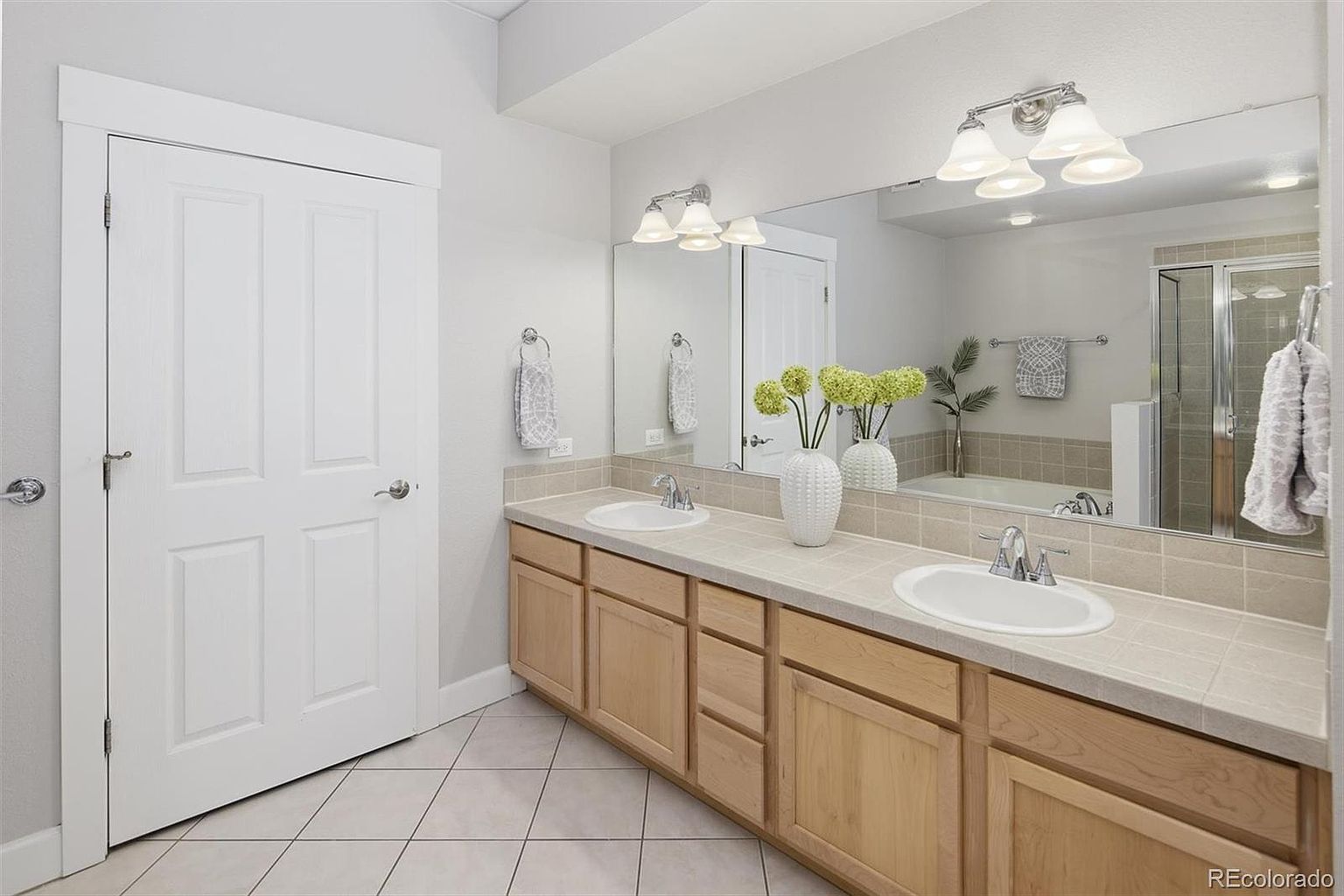 This spacious primary bathroom features a dual-sink vanity with light wood cabinetry and neutral tile countertops. A large wall-to-wall mirror reflects the bright, clean space, which includes a separate shower area and a neutral color palette. The room is well-lit by two vanity light fixtures, creating a functional and inviting atmosphere.