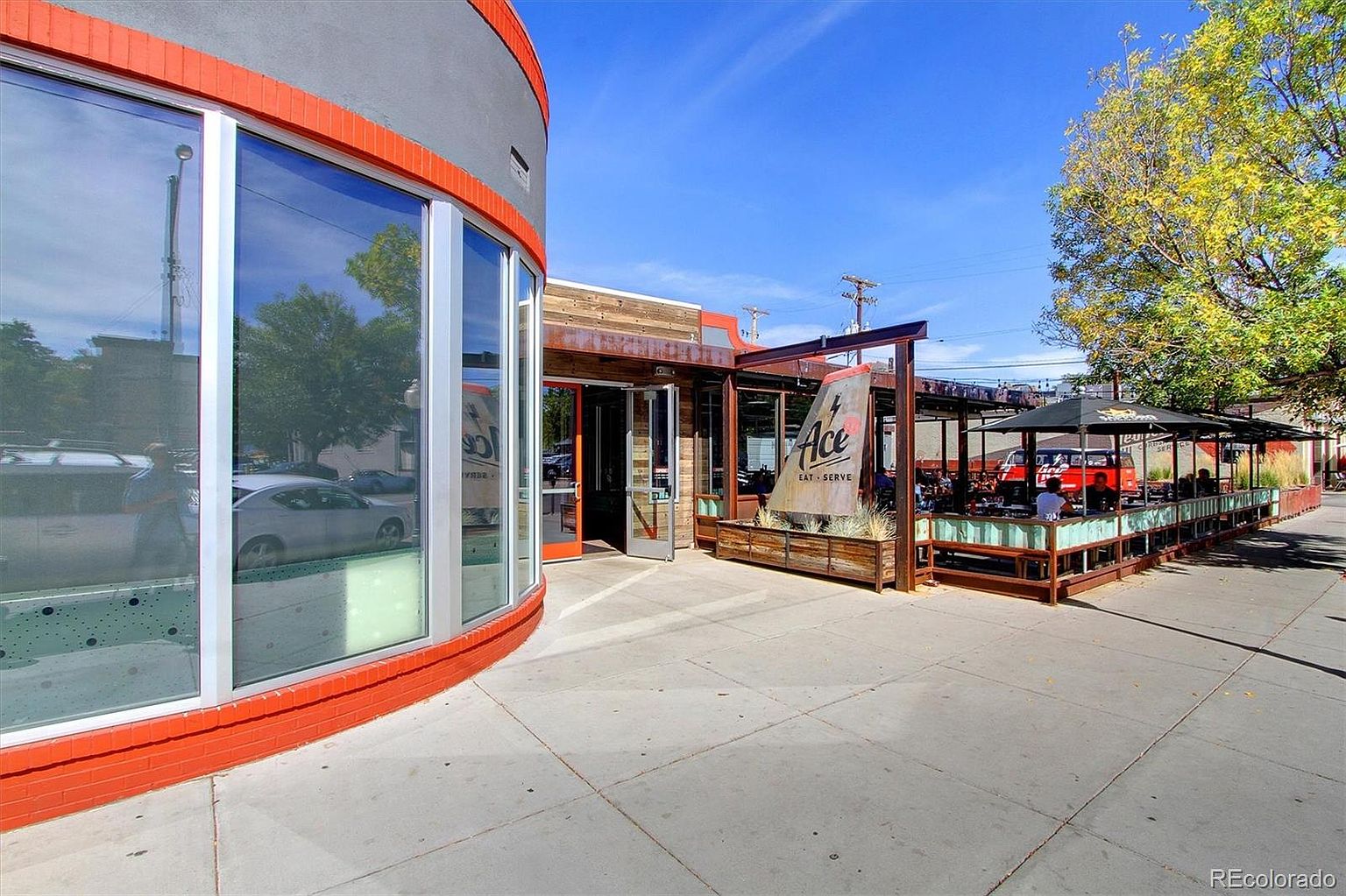 This image captures the exterior of a vibrant, modern restaurant featuring a curved brick-accented facade and a spacious outdoor patio dining area. The scene is bathed in bright, clear daylight, highlighting the industrial-chic aesthetic with wood paneling and metal structures. The perspective is a wide-angle street-level view, showcasing the inviting atmosphere of the establishment's entrance and seating area.