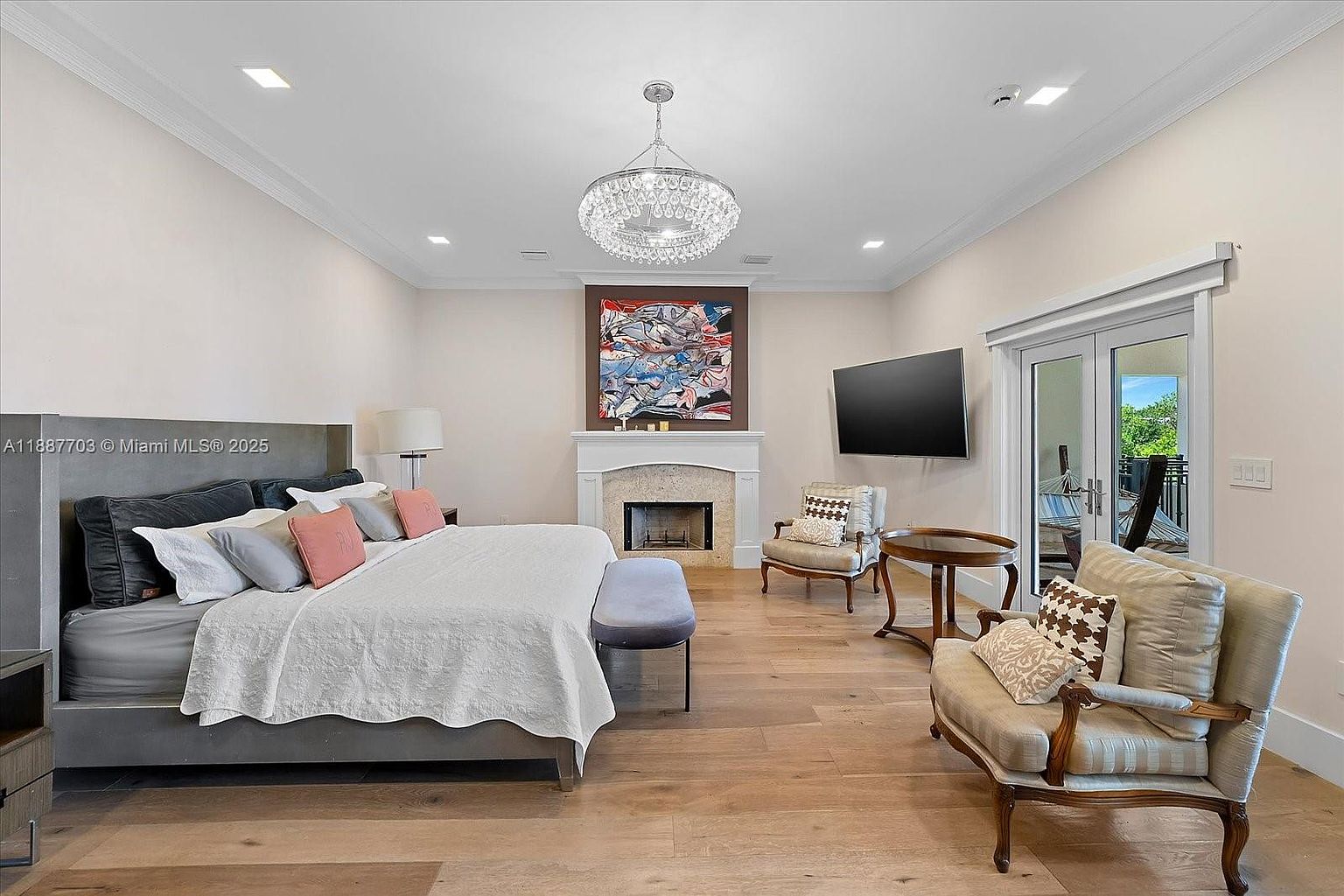 This is a primary bedroom featuring a large bed with a gray headboard and white bedding, complemented by colorful pillows. The room includes a fireplace with an abstract painting above it, a television mounted on the wall, and two armchairs with a small table between them. French doors lead to an outdoor balcony, and the hardwood floors add warmth to the space.