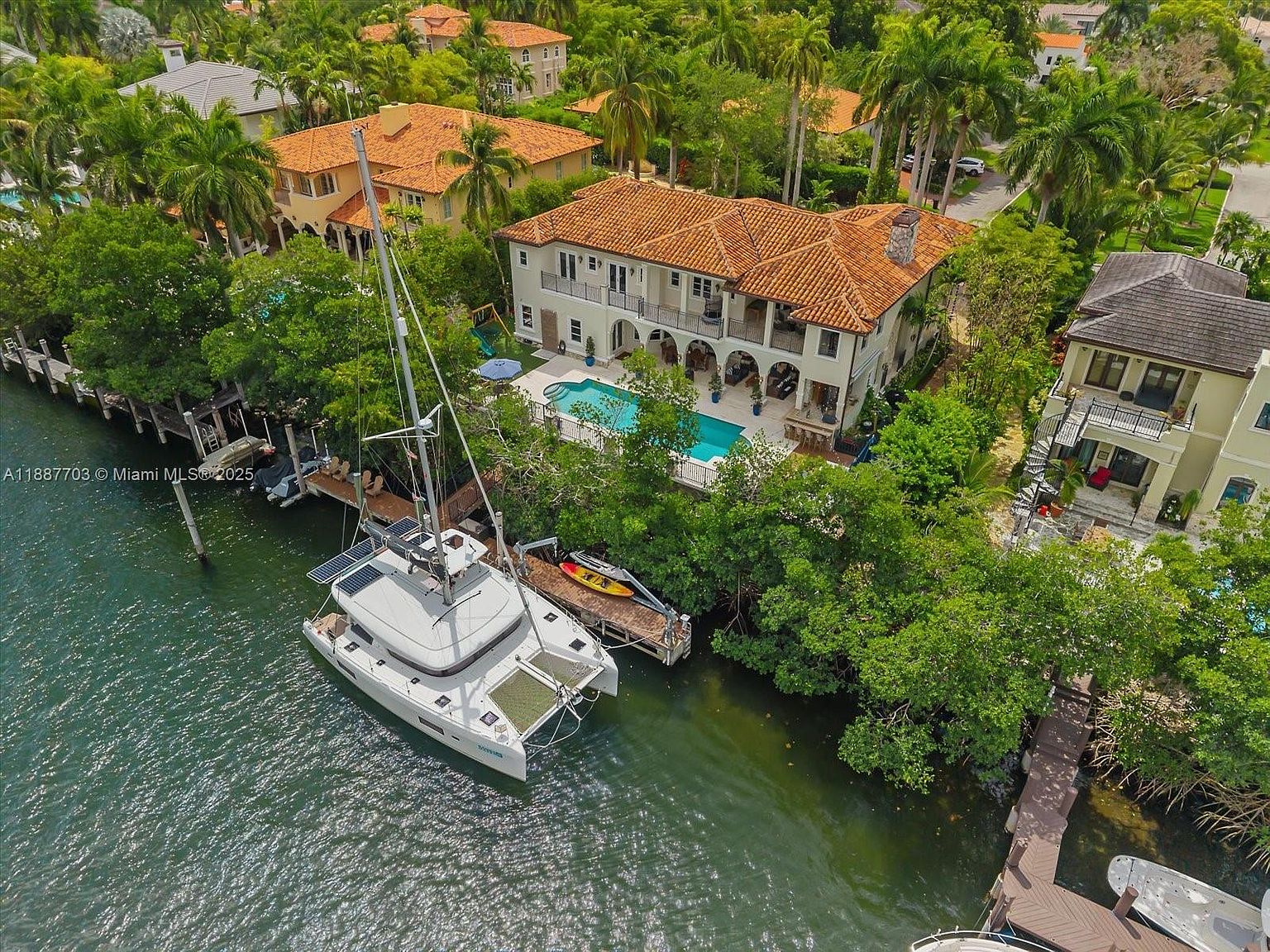 This aerial shot showcases a luxurious waterfront property featuring a Mediterranean-style house with a terracotta tile roof, a private pool, and a boat docked at a private pier. Lush greenery surrounds the property, providing privacy and enhancing the overall appeal. The image highlights the property's prime location and upscale amenities.