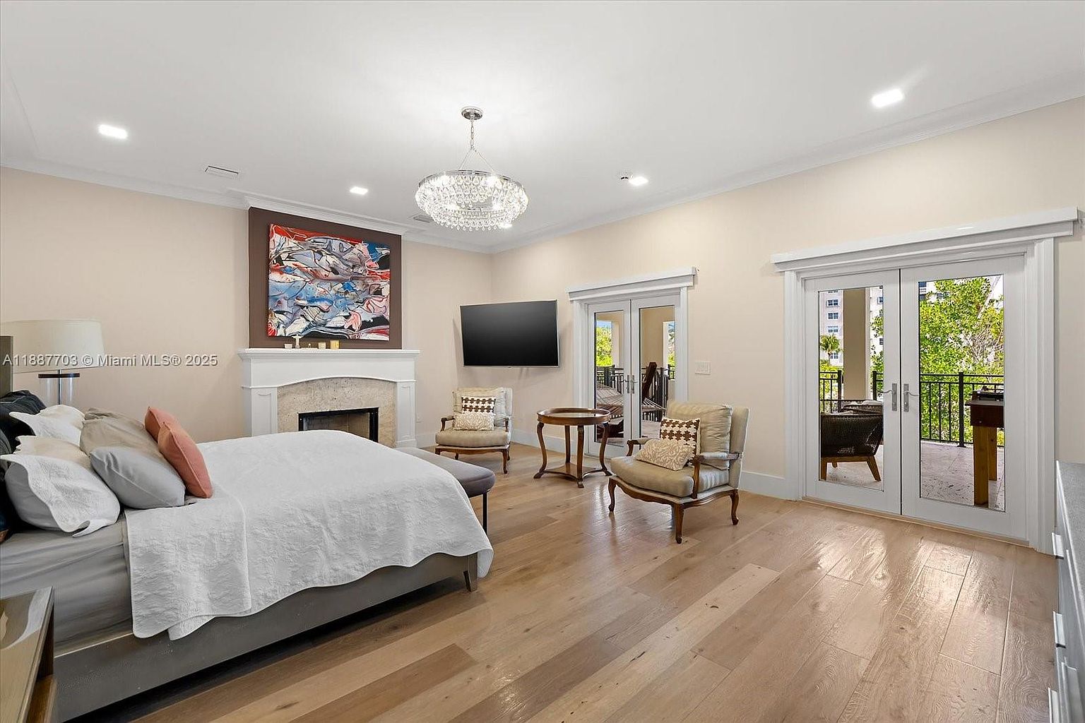 This is a primary bedroom featuring a bed with white linens, a fireplace with a decorative mantle, and a large piece of artwork above the fireplace. There is a television mounted on the wall, two chairs with a small table between them, and two sets of French doors leading to a balcony. The room has light-colored walls and hardwood floors, creating a bright and airy atmosphere.