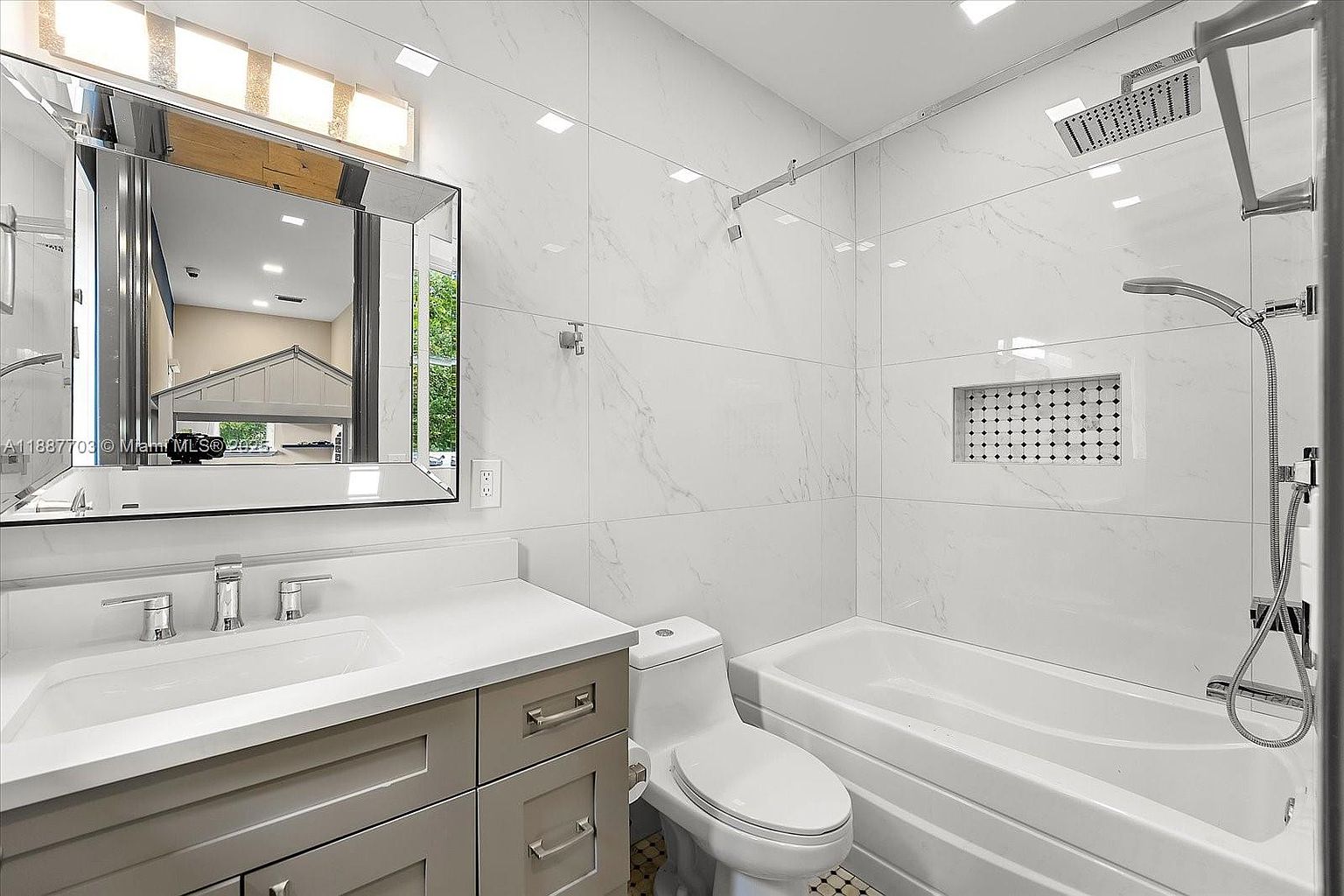 This is a well-lit bathroom featuring white marble-style tiling on the walls and a combination shower and bathtub. The vanity has a light gray cabinet with a white countertop and a modern sink. A toilet is positioned next to the vanity, and a large mirror hangs above the sink, reflecting the interior of the house.