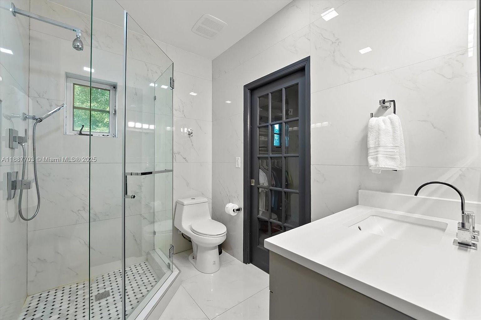 This is a primary bathroom featuring a glass-enclosed shower with a rainfall showerhead and a small window. The walls and floors are covered in white marble-like tiles, creating a clean and luxurious feel. A modern vanity with a white countertop and gray cabinetry is visible, along with a toilet and a dark-framed door, adding a touch of contrast to the space.