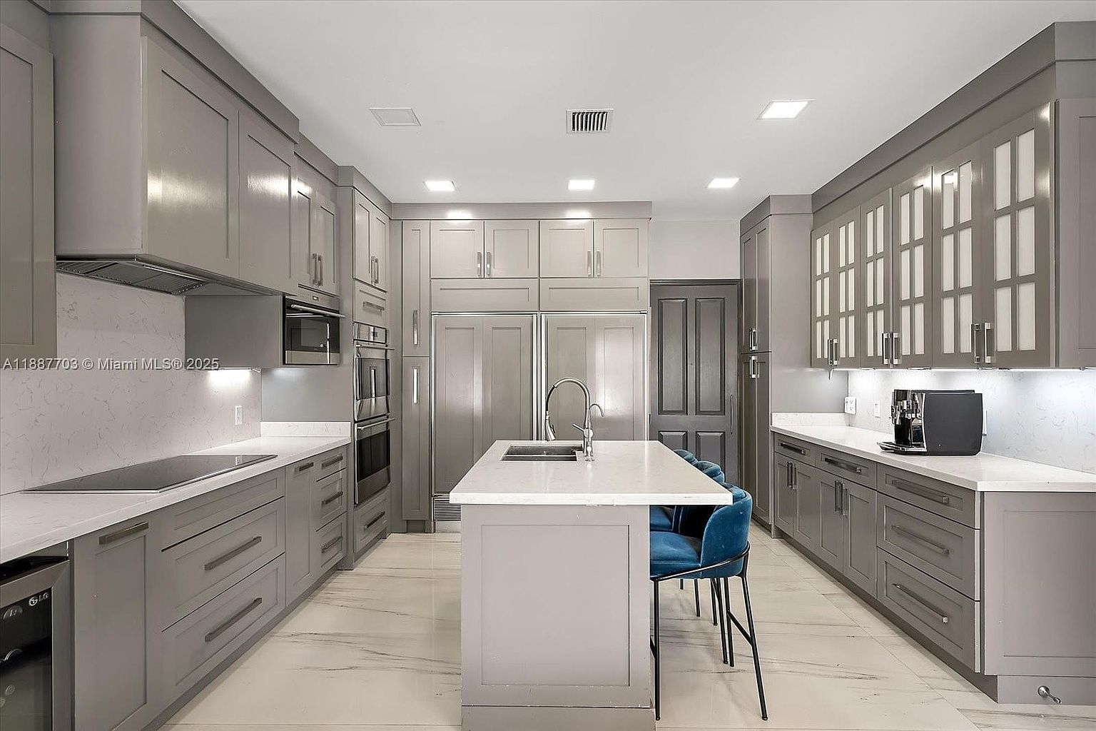 This is a modern kitchen with gray cabinetry, stainless steel appliances, and a large island with a white countertop. The kitchen features a clean and contemporary design, with recessed lighting and a marble floor. The island has blue bar stools, and there is a coffee machine on the counter.