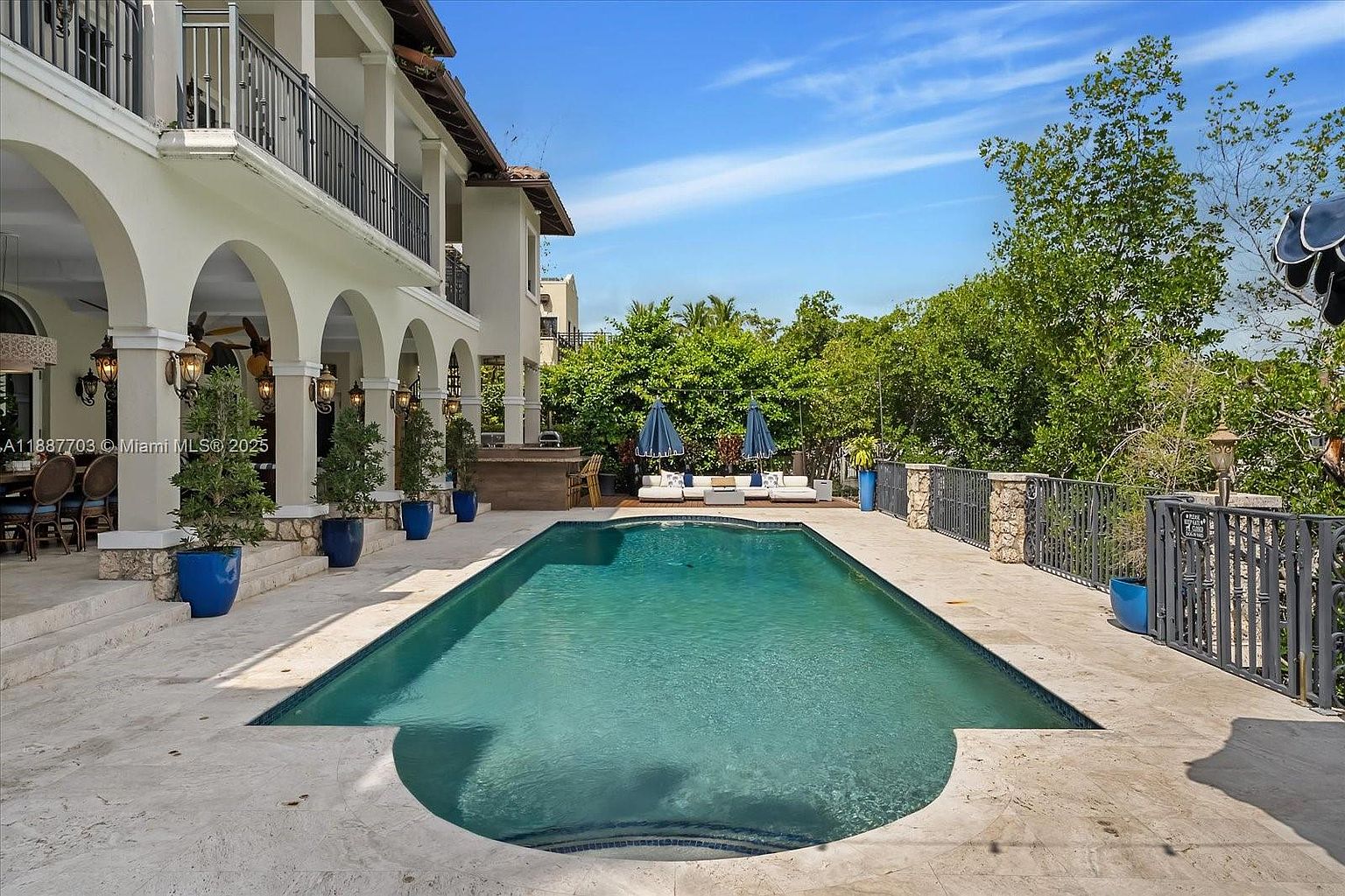 This image showcases a luxurious backyard pool area. The rectangular pool is surrounded by a light-colored stone patio, with elegant potted plants and outdoor furniture adding to the ambiance. A covered patio with arched columns is attached to the house, and a decorative fence borders the property, creating a private and upscale outdoor living space.