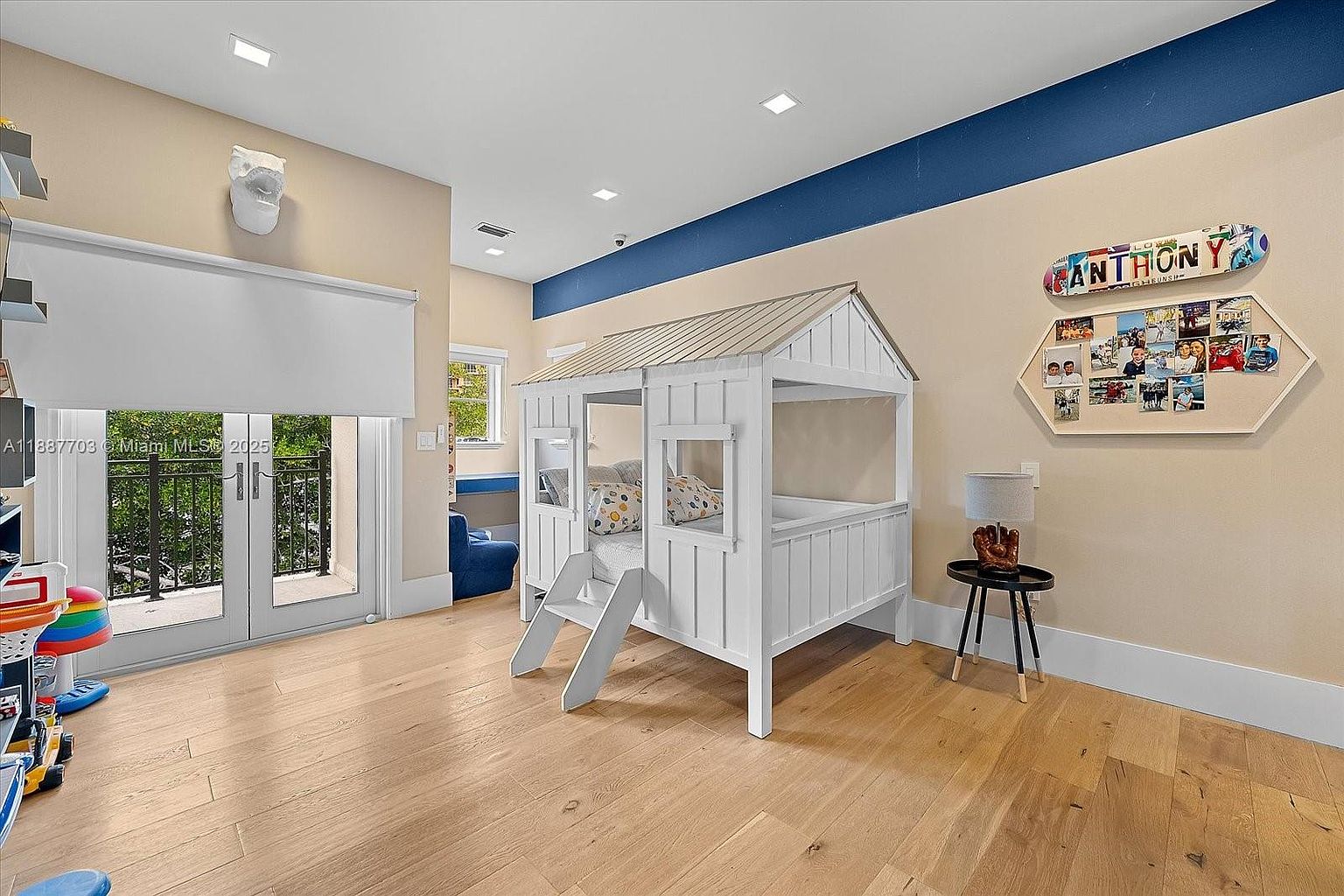 This is a bright and playful bedroom featuring a unique house-shaped bed with a white frame and a light-colored roof. The room has light hardwood floors and neutral-toned walls with a blue accent stripe near the ceiling. A glass door leads to a balcony, and a small side table with a lamp and a baseball glove adds a touch of personality.