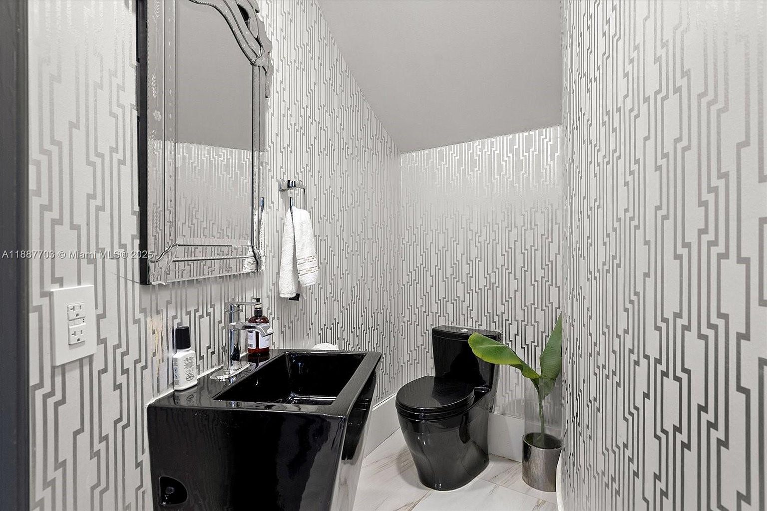 This is a modern guest bathroom featuring a black pedestal sink and toilet, complemented by a striking geometric wallpaper. A decorative mirror hangs above the sink, and a small plant adds a touch of nature to the space. The overall design is sleek and stylish, creating a sophisticated and contemporary feel.