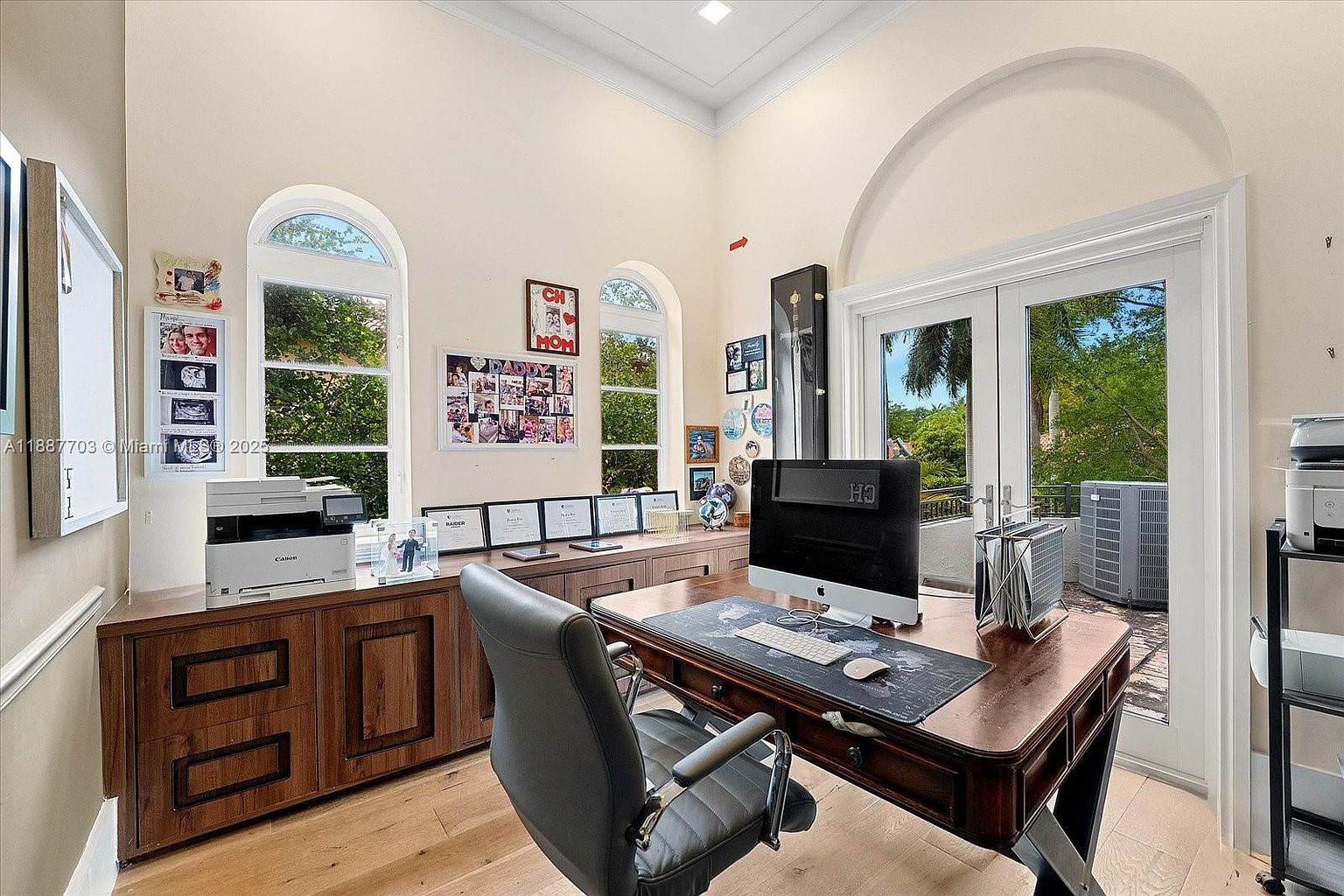 This is an interior shot of a home office featuring a large wooden desk with a computer setup, including a monitor, keyboard, and mouse. The room has a high ceiling and arched windows that provide natural light. A wooden cabinet runs along the wall, and a comfortable office chair is positioned in front of the desk, creating a professional and inviting workspace.