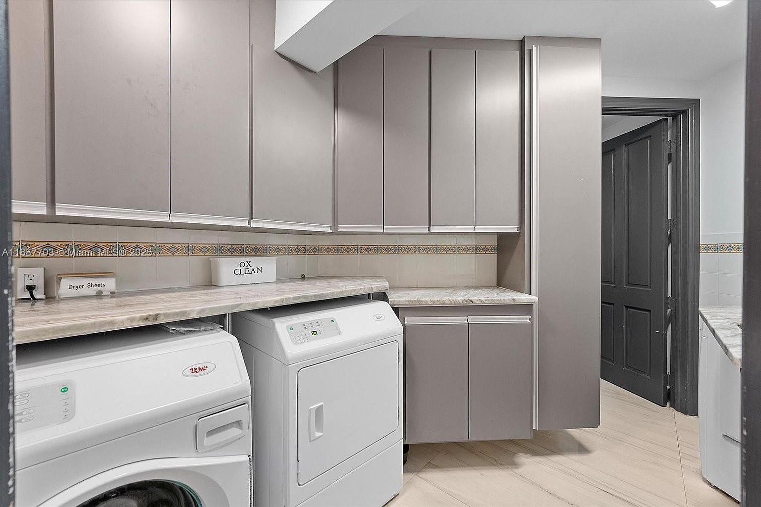 This is an interior shot of a laundry room featuring a white washer and dryer set under a light-colored countertop. Above the countertop are gray cabinets, and a doorway is visible on the right side of the frame. The room has a clean and organized appearance, suggesting a functional and well-maintained space.