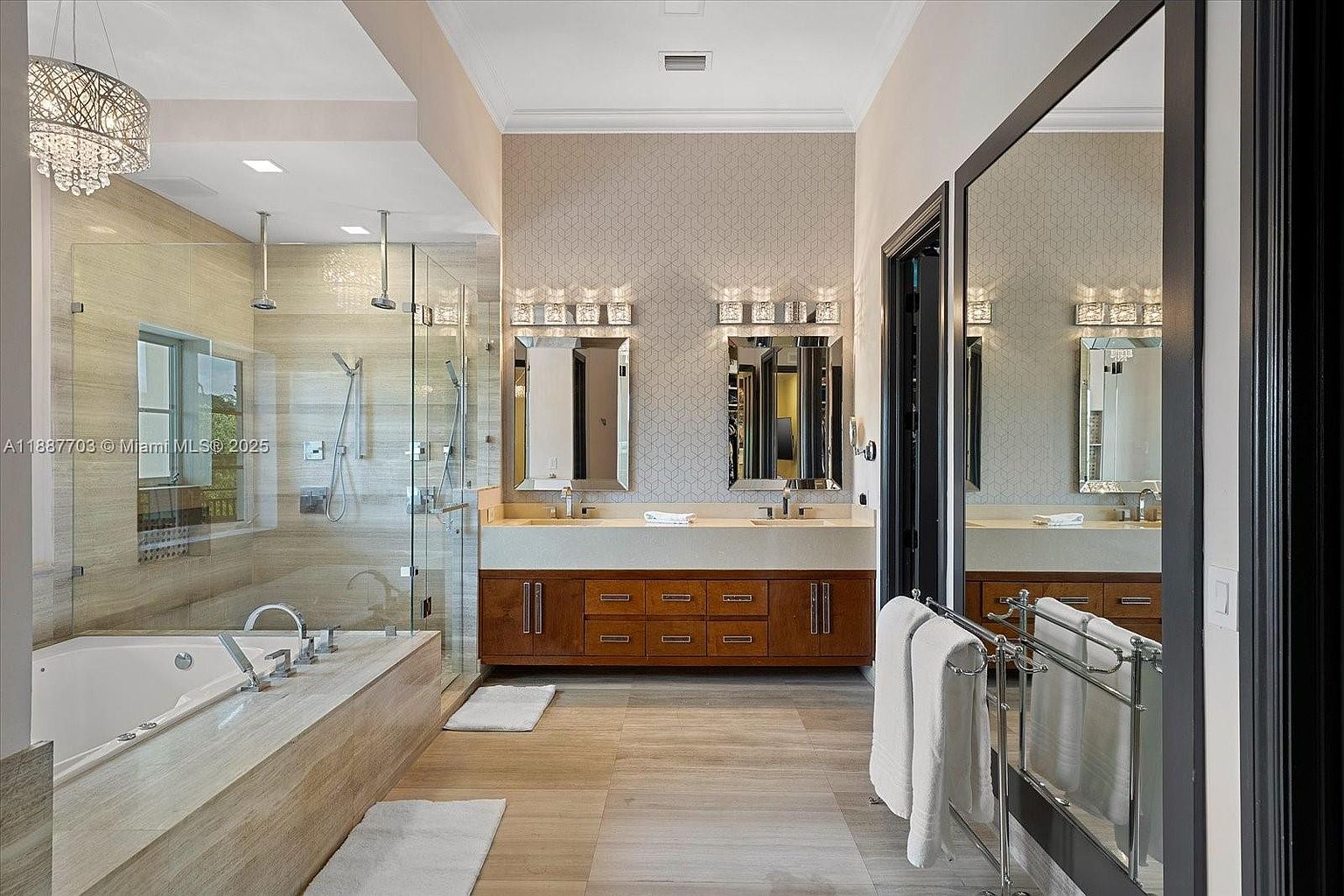 This is a luxurious primary bathroom featuring a large soaking tub and a separate glass-enclosed shower with dual shower heads. The double vanity has wood cabinetry, light countertops, and decorative mirrors with sconces. The flooring is light-colored tile, and the overall design is modern and elegant.