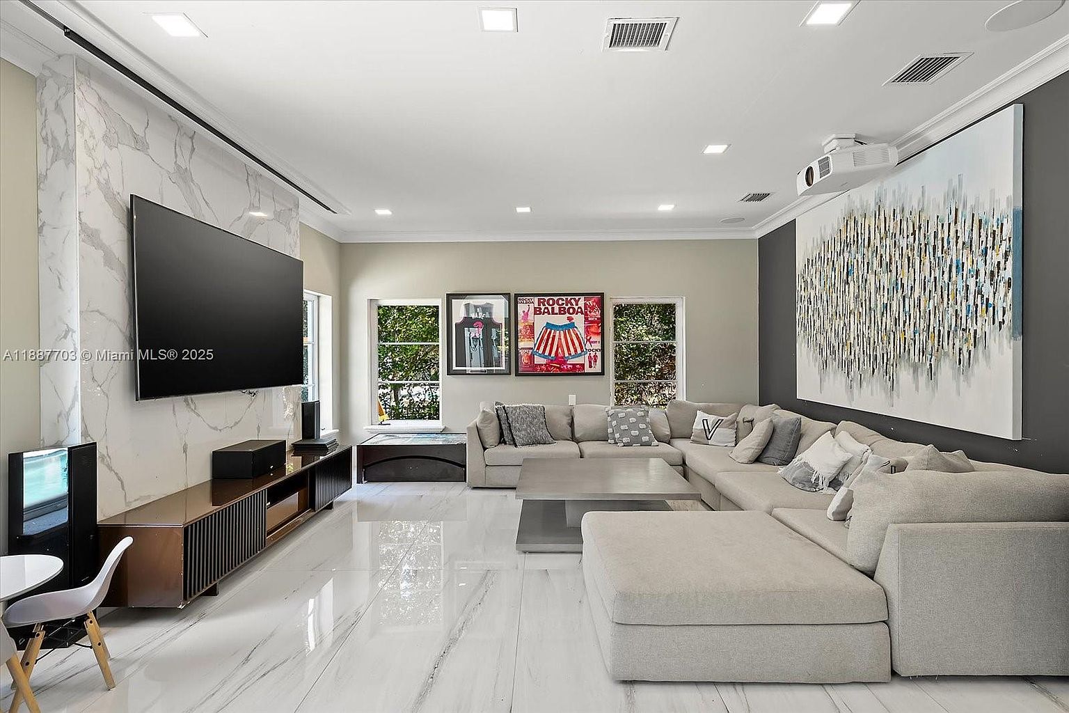 This is an interior shot of a modern living room featuring a large sectional sofa, a sleek entertainment center with a large flat-screen TV, and marble flooring. The room is well-lit with recessed lighting and natural light from the windows, which are adorned with framed art. The overall impression is one of spaciousness and contemporary elegance.