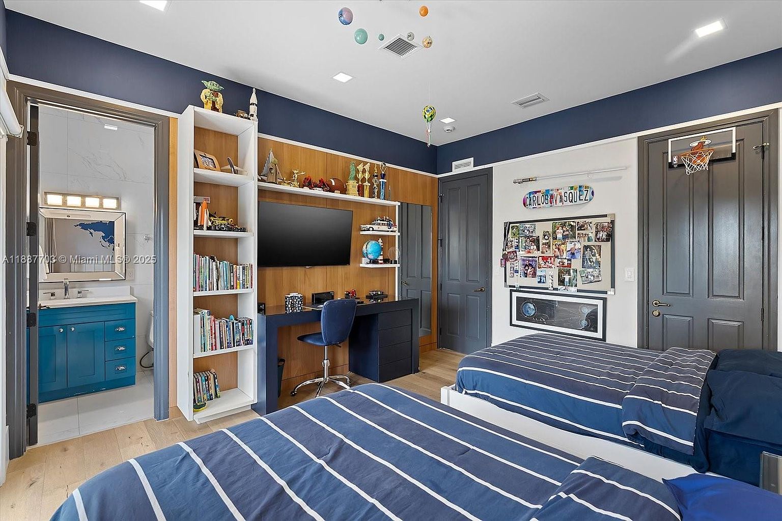 This is a well-decorated bedroom featuring two twin beds with blue and white striped bedding. The room includes a built-in desk area with shelving and a TV, creating a functional space for work and entertainment. The color scheme is predominantly blue and white, giving the room a clean and modern feel.