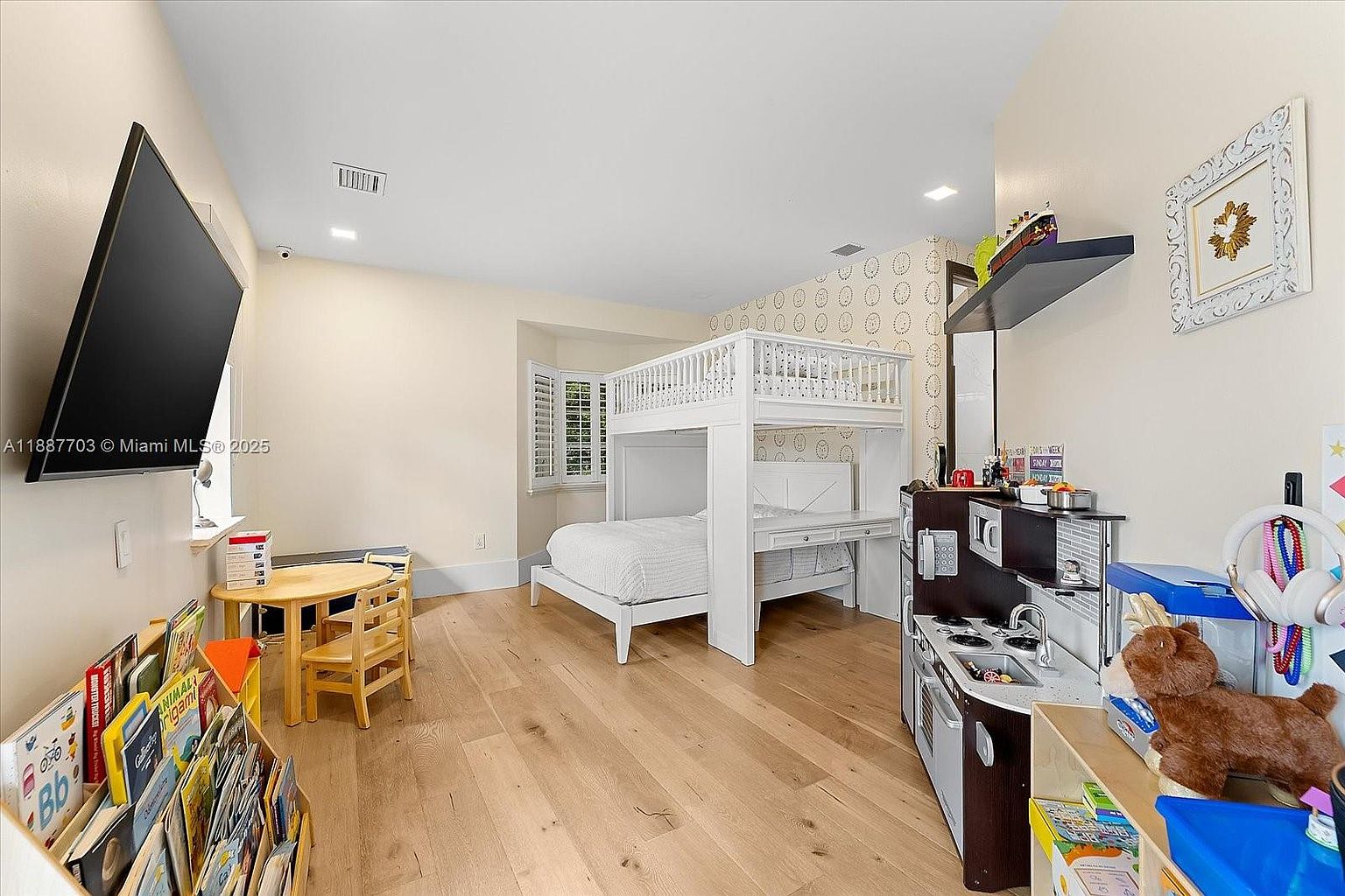 This is a well-organized children's bedroom featuring a white bunk bed with a built-in desk, a wooden table with chairs, and a bookshelf filled with books. A play kitchen set adds a playful touch to the room. The room has light hardwood floors and neutral-colored walls, creating a bright and inviting atmosphere.