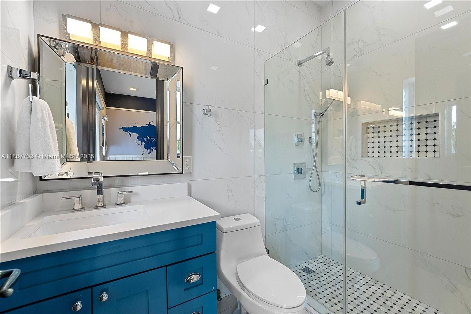 This is a well-lit primary bathroom featuring a blue vanity with a white countertop and a modern sink. A large mirror with integrated lighting hangs above the vanity. The bathroom also includes a toilet and a glass-enclosed shower with white marble-like tiling and black and white mosaic floor tiles, creating a clean and luxurious feel.