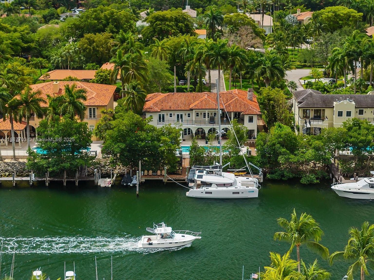 This aerial shot showcases a luxurious waterfront property with a Mediterranean-style house featuring a red tile roof and white facade. A large catamaran is docked at the property's private pier, and a smaller boat is seen cruising on the waterway. Lush tropical landscaping surrounds the house, enhancing its appeal and privacy.
