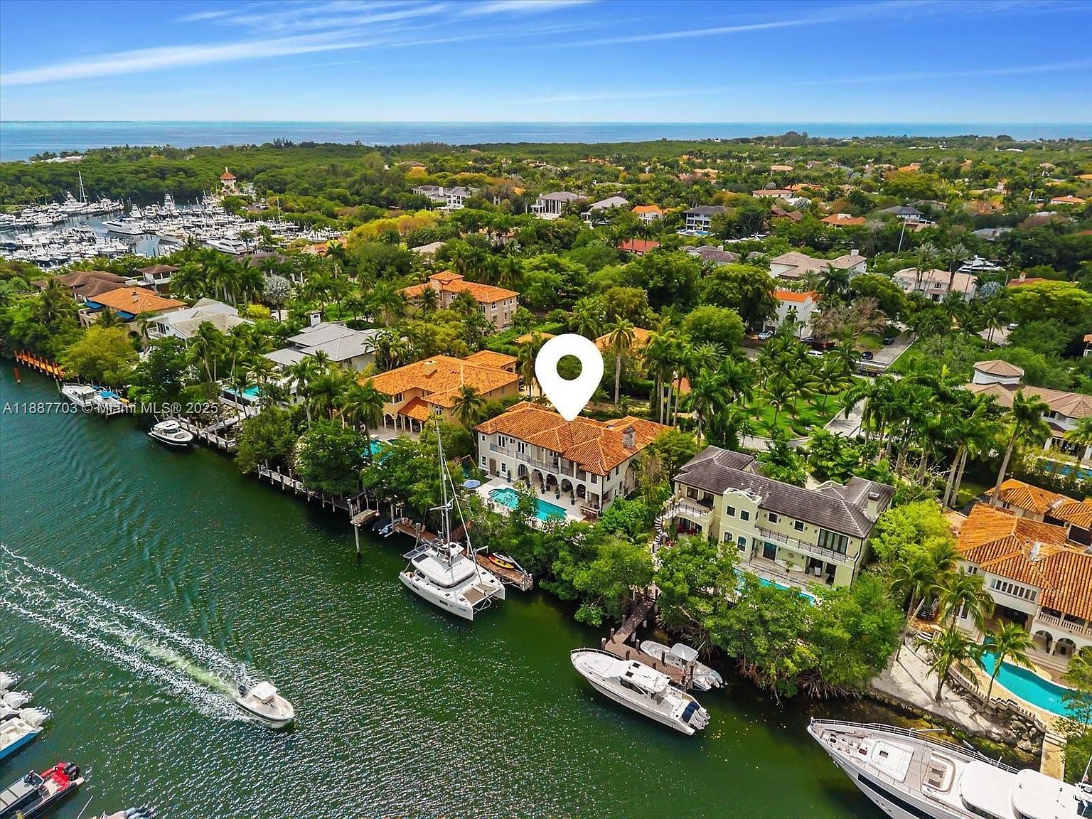 This aerial view showcases a luxurious waterfront property with a Mediterranean-style villa featuring a terracotta tile roof and manicured landscaping. The property includes a private dock with boats, a swimming pool, and lush greenery, offering a serene and upscale living environment. The perspective is from above, looking down at the property and the surrounding waterfront.