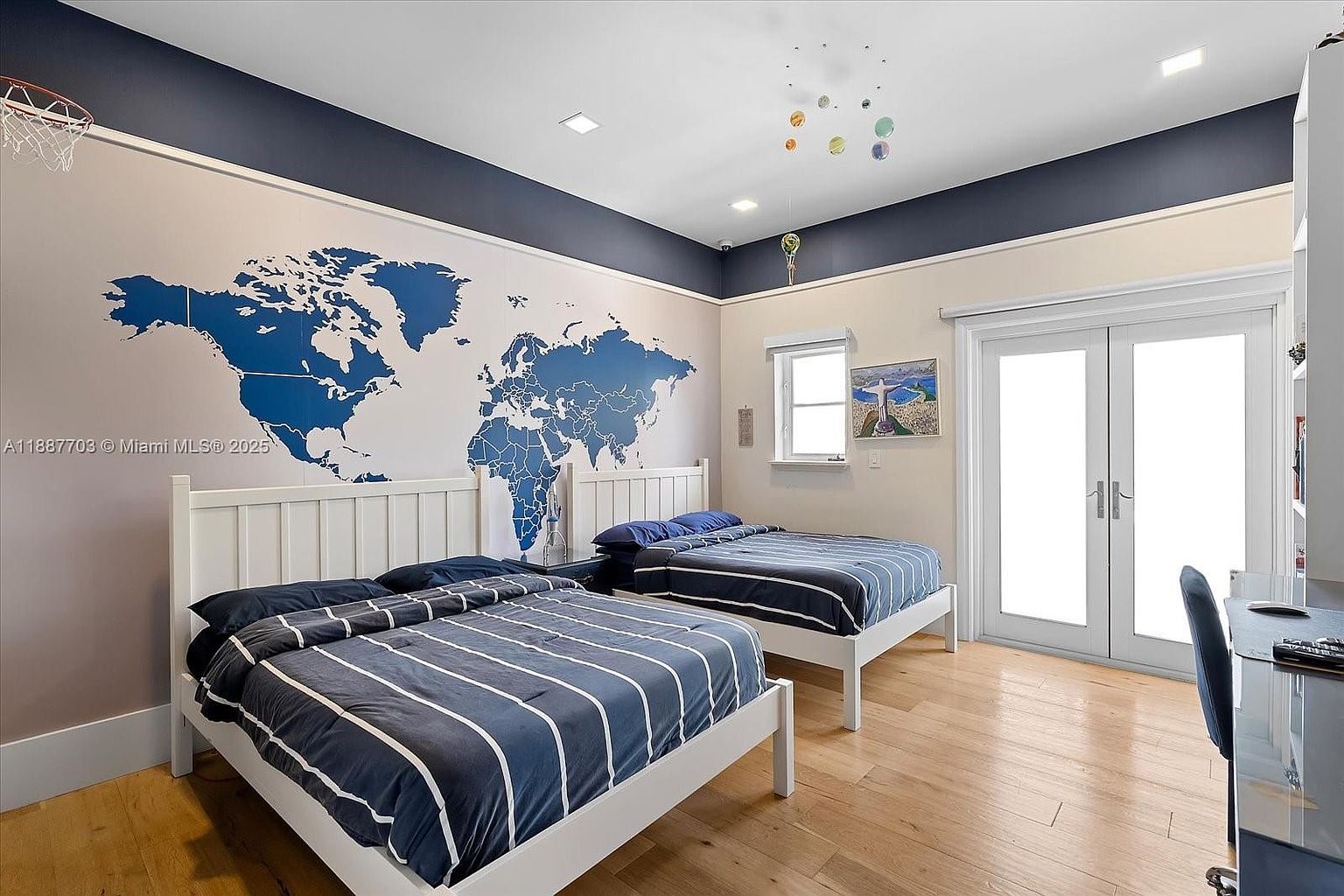 This is a well-lit guest bedroom featuring two twin beds with white frames and blue striped bedding. A large world map mural adorns the wall, adding a playful and educational touch. The room has hardwood floors and a door leading to an outdoor area, creating a bright and inviting space.