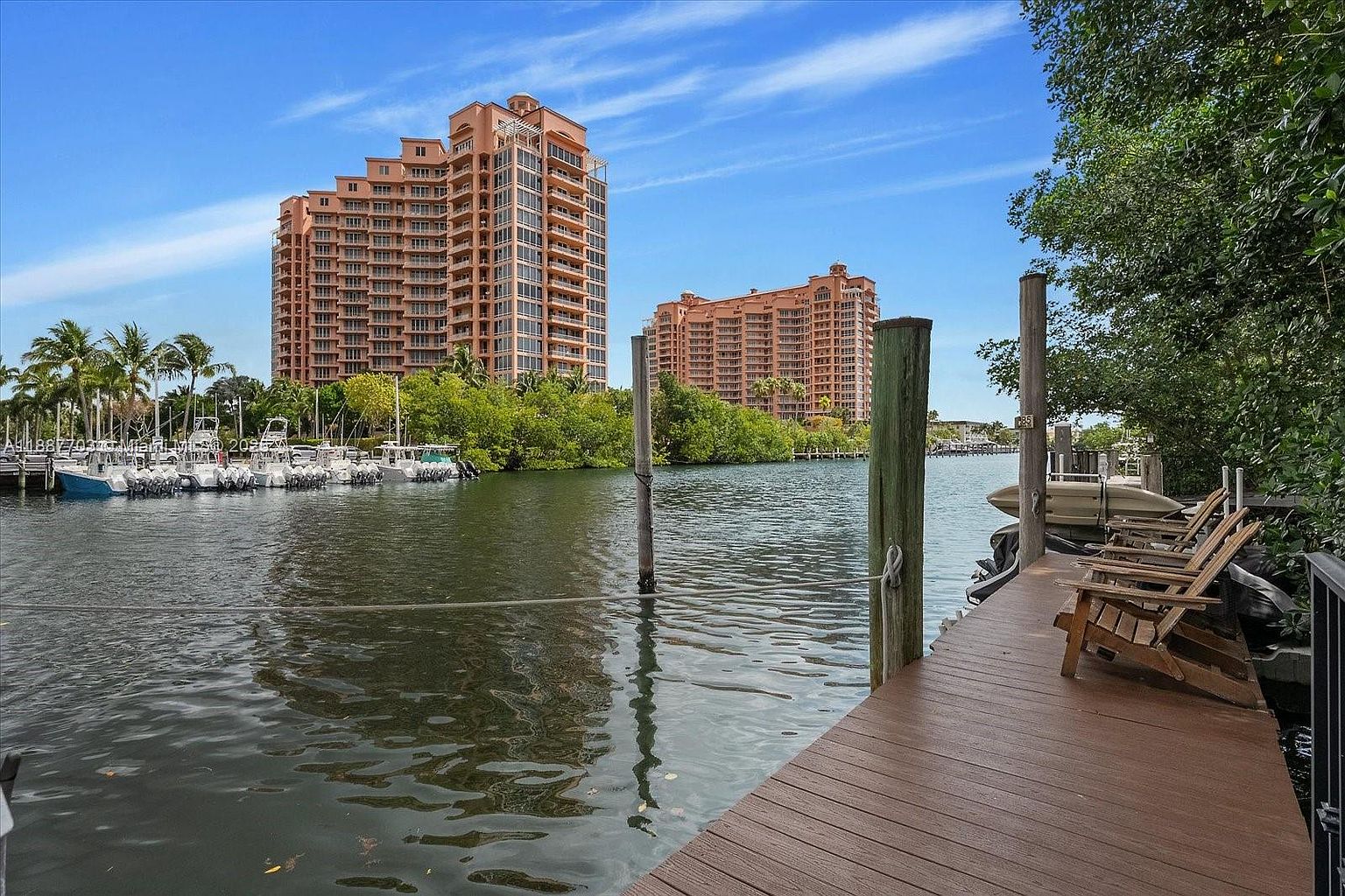 This image showcases a waterfront patio or deck with wooden flooring and chairs, offering a serene view of the water and nearby buildings. The scene includes lush greenery, creating a tranquil and inviting outdoor space. The presence of boats and the waterfront setting enhance the property's appeal, suggesting a luxurious and relaxing lifestyle.