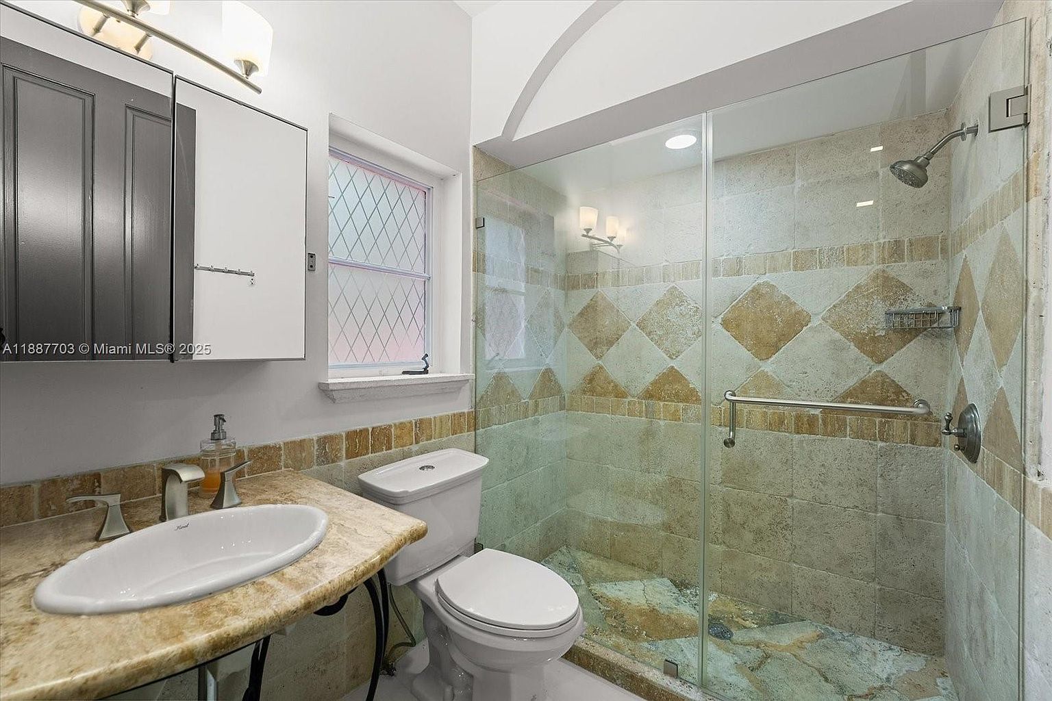 This is a bathroom featuring a marble countertop with a white sink, a toilet, and a glass-enclosed shower. The shower has decorative tile work with diamond patterns and a built-in shower head. The overall impression is clean and functional, with a touch of elegance from the marble and tile.