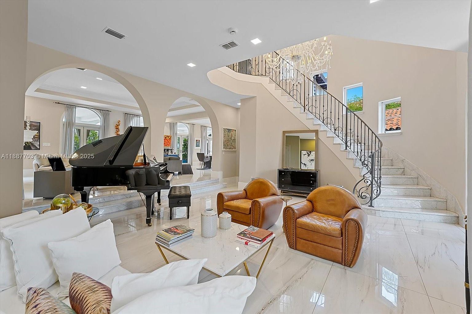 This is a luxurious living room featuring a grand piano, elegant staircase with wrought iron railings, and high ceilings. The room is decorated with modern furniture, including leather armchairs and a white sofa with decorative pillows. The marble flooring and chandelier add to the opulent feel of the space, creating an inviting and sophisticated atmosphere.