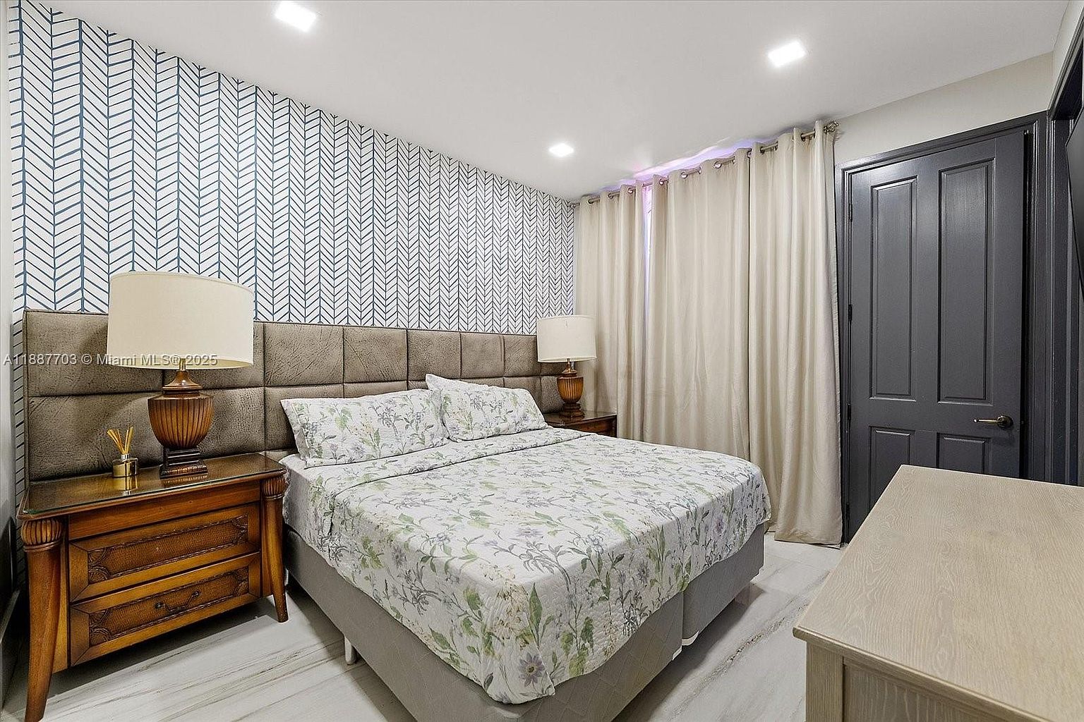This is a primary bedroom featuring a bed with a floral patterned comforter and pillows, flanked by wooden nightstands with lamps. The headboard is upholstered in a neutral tone, and the wall behind the bed is covered in a geometric patterned wallpaper. The room also includes neutral-toned curtains and a dark-colored door, creating a serene and inviting atmosphere.