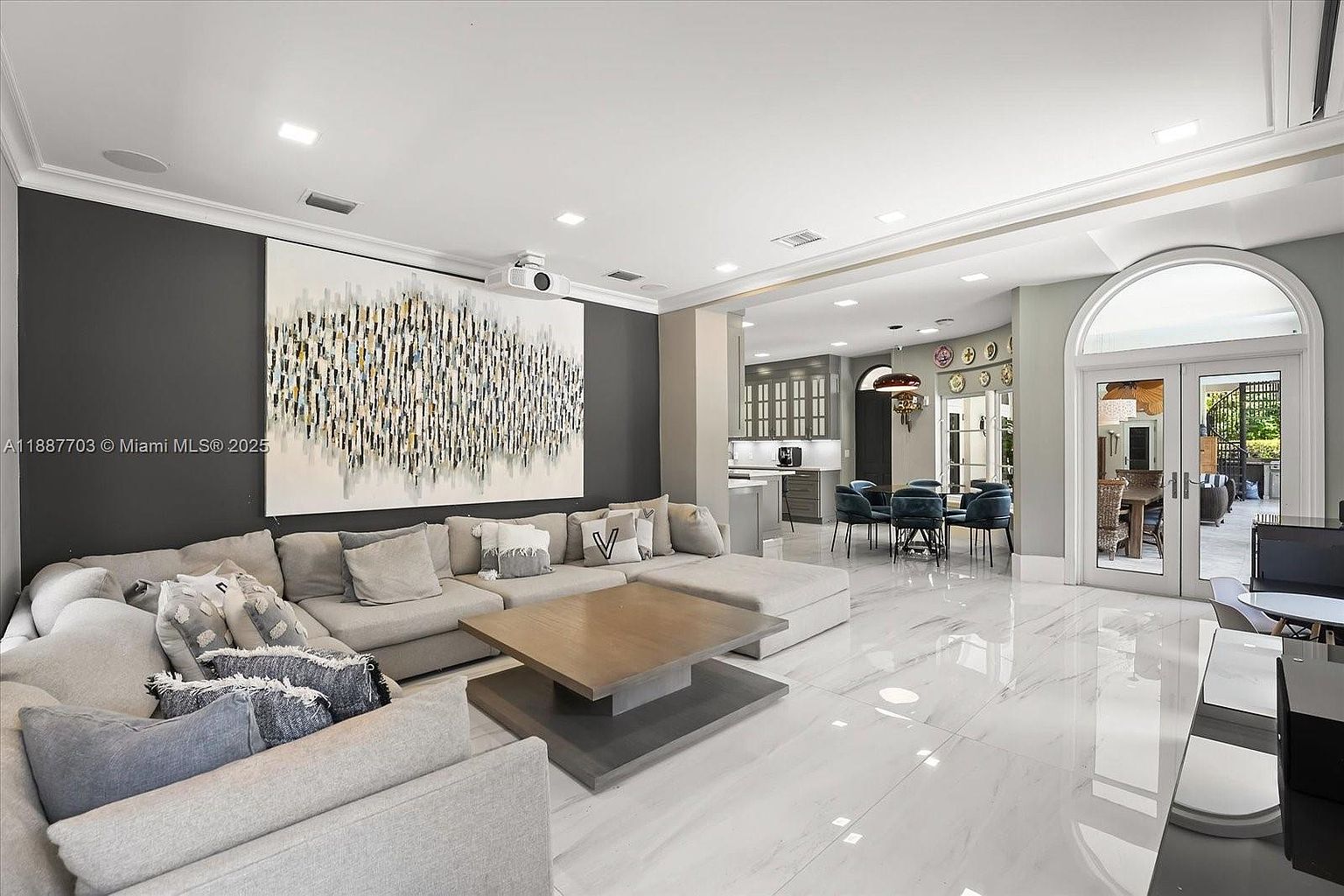 This is a spacious and luxurious living room featuring a large sectional sofa, a modern coffee table, and a striking piece of abstract art on the wall. The room boasts high ceilings, recessed lighting, and polished marble floors, creating an open and elegant atmosphere. The open floor plan seamlessly connects to the dining area and kitchen, enhancing the sense of flow and connectivity.