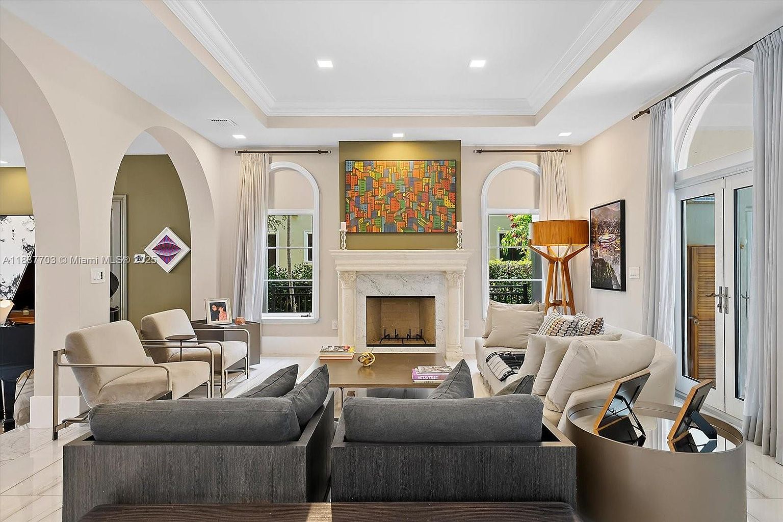 This is an interior shot of a well-lit living room featuring two gray sofas, two beige armchairs, and a marble fireplace with a colorful painting above it. The room has high ceilings with recessed lighting, arched windows with sheer curtains, and a modern, elegant design. A round, reflective coffee table adds a touch of sophistication.