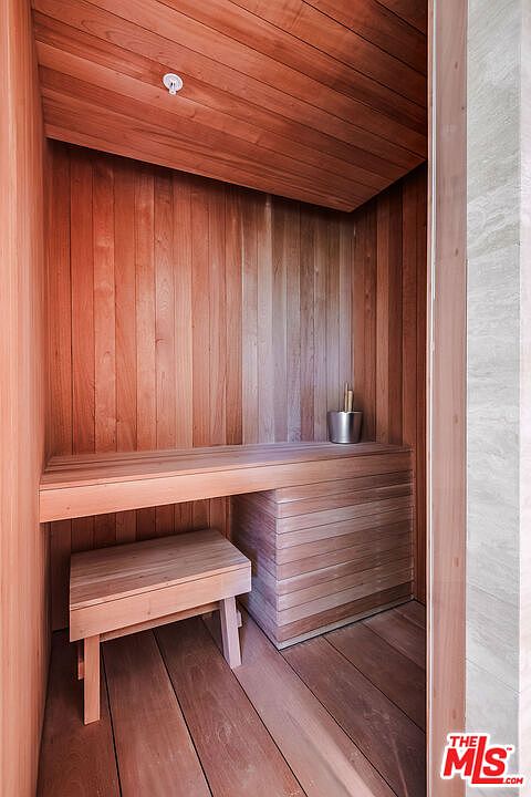 This image showcases a compact, custom-built cedar sauna, likely located within a primary bathroom suite. The space features warm, natural wood paneling throughout, a built-in bench, and a small stool, creating a serene and spa-like atmosphere. The perspective is framed from the doorway, highlighting the craftsmanship and inviting, tranquil nature of this wellness amenity.