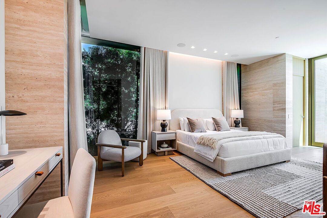 This sophisticated primary bedroom features a minimalist design with warm, natural wood flooring and elegant travertine-style wall accents. A plush, upholstered bed serves as the focal point, complemented by a cozy reading nook with a modern armchair and floor-to-ceiling windows that invite lush greenery inside. The space is bathed in soft, natural light, creating a serene and luxurious atmosphere perfect for a high-end residence.