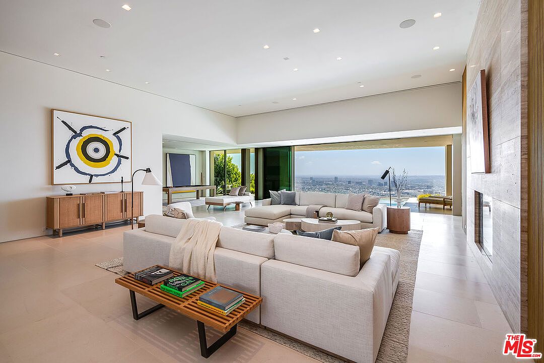 This expansive, ultra-modern living room features a minimalist aesthetic with high ceilings, neutral tones, and a seamless transition to an outdoor terrace overlooking a sprawling city skyline. The space is anchored by a large, plush sectional sofa, a wooden slat bench, and a striking abstract art piece, all illuminated by recessed lighting and abundant natural light. The open-concept design emphasizes luxury and architectural sophistication, creating an airy and inviting atmosphere.