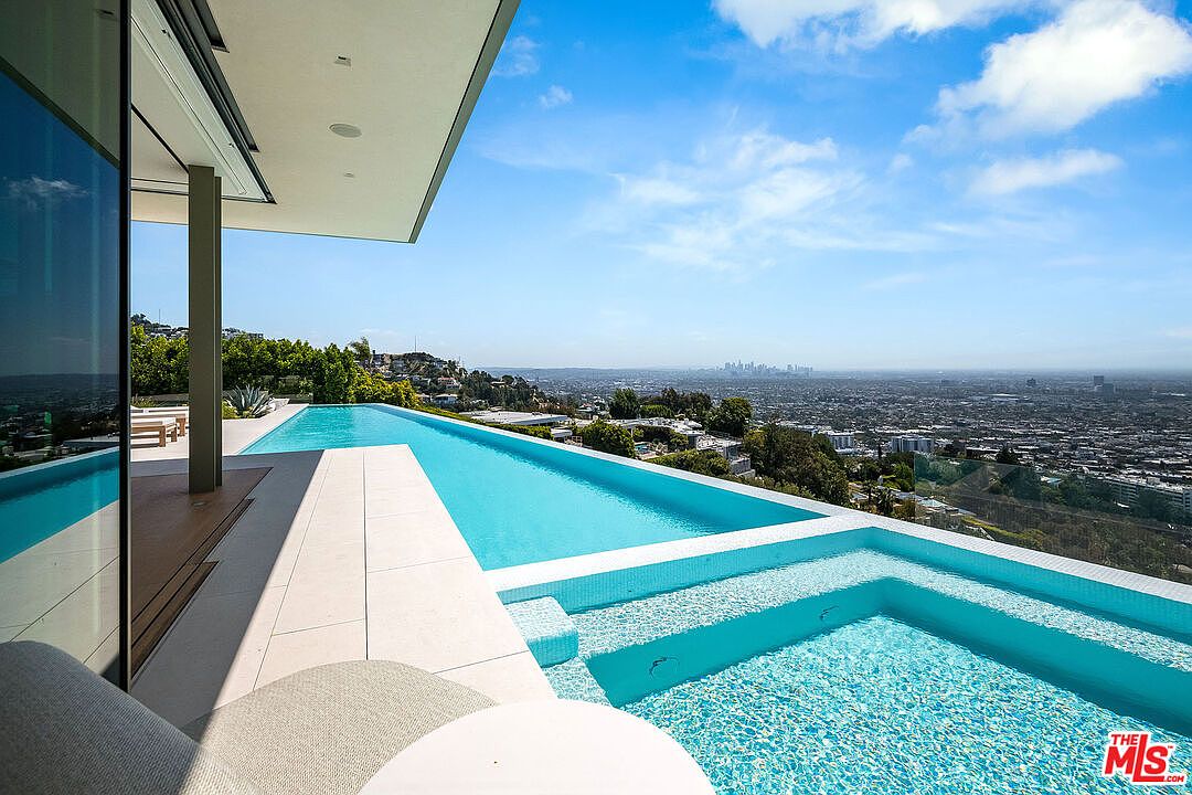 This stunning infinity-edge pool and integrated spa offer a breathtaking panoramic view of the sprawling city skyline below. The sleek, modern design features clean lines and light-colored tiling that perfectly complements the bright blue water and clear sky. The perspective from the adjacent patio provides a luxurious, high-end feel, emphasizing the seamless transition between the home's architecture and the expansive outdoor horizon.