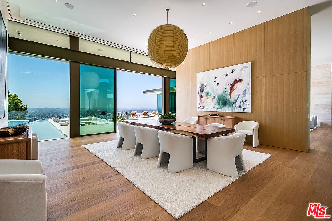 This sophisticated dining room features a long, natural-edge wooden table surrounded by six modern, white upholstered chairs, set atop a plush cream rug. A large, spherical paper pendant light hangs above, while a vibrant abstract painting adds a pop of color to the warm wood-paneled wall. The room seamlessly transitions to an outdoor terrace and infinity pool through floor-to-ceiling glass sliding doors, offering a breathtaking panoramic view of the city below.