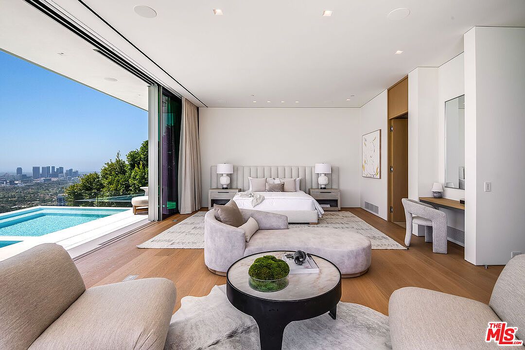 This luxurious primary bedroom features a contemporary design with a seamless indoor-outdoor flow, highlighted by floor-to-ceiling glass walls that open directly to a private pool deck overlooking a sprawling city skyline. The room is furnished with a plush, light-toned bed, a curved chaise lounge, and a sleek vanity area, all set against a backdrop of neutral, minimalist decor. The perspective is captured from the foot of the bed, emphasizing the expansive, open-concept layout and the breathtaking panoramic views.