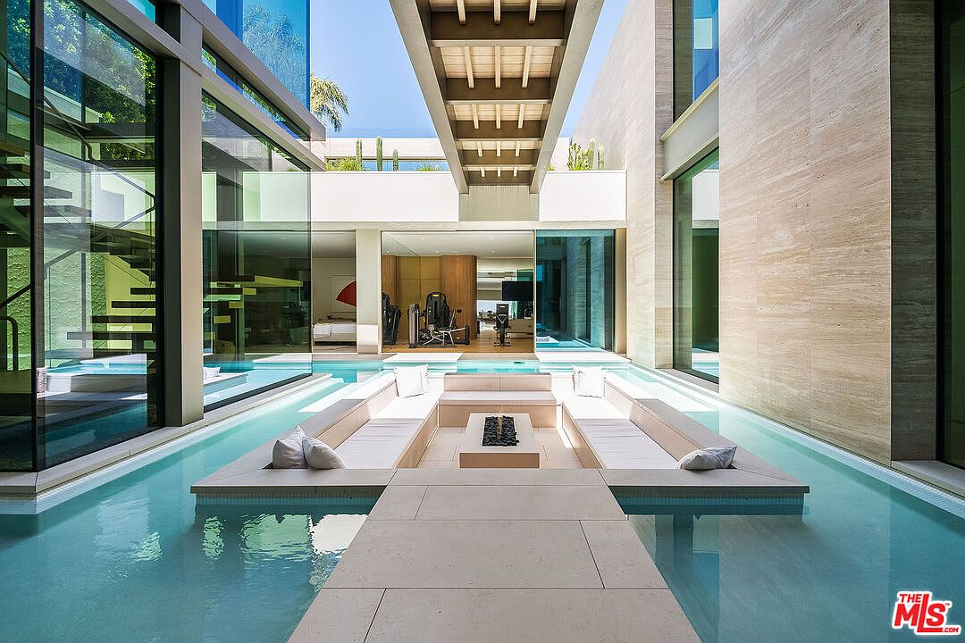 This stunning architectural courtyard features a sunken seating area surrounded by a tranquil, crystal-clear reflecting pool. The space is framed by floor-to-ceiling glass walls and sleek stone surfaces, creating a seamless blend of indoor and outdoor luxury living. A modern overhead bridge structure adds a dramatic geometric element to this sophisticated, resort-style oasis.