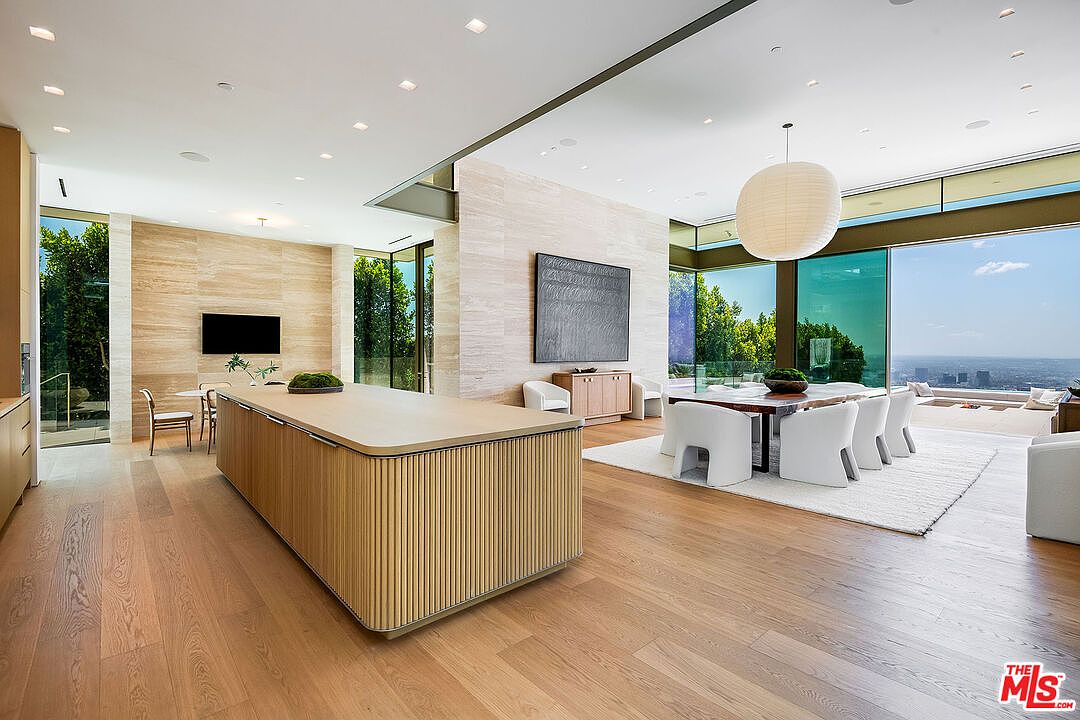 This expansive, open-concept kitchen and dining area showcases a sophisticated modern aesthetic with light wood flooring and textured stone walls. A large, fluted-wood kitchen island serves as the centerpiece, complemented by a minimalist dining set and floor-to-ceiling glass walls that offer panoramic city views. The space is bathed in natural light, creating an airy, luxurious atmosphere perfect for high-end entertaining.