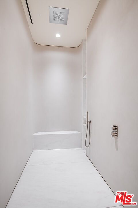 This minimalist, high-end walk-in shower features seamless white stone surfaces and a built-in bench, creating a serene and spa-like atmosphere. The space is equipped with a modern ceiling-mounted rainfall showerhead and a sleek handheld sprayer, emphasizing a clean, contemporary aesthetic. The perspective is a straight-on, eye-level shot that highlights the architectural simplicity and luxurious materials of the shower enclosure.