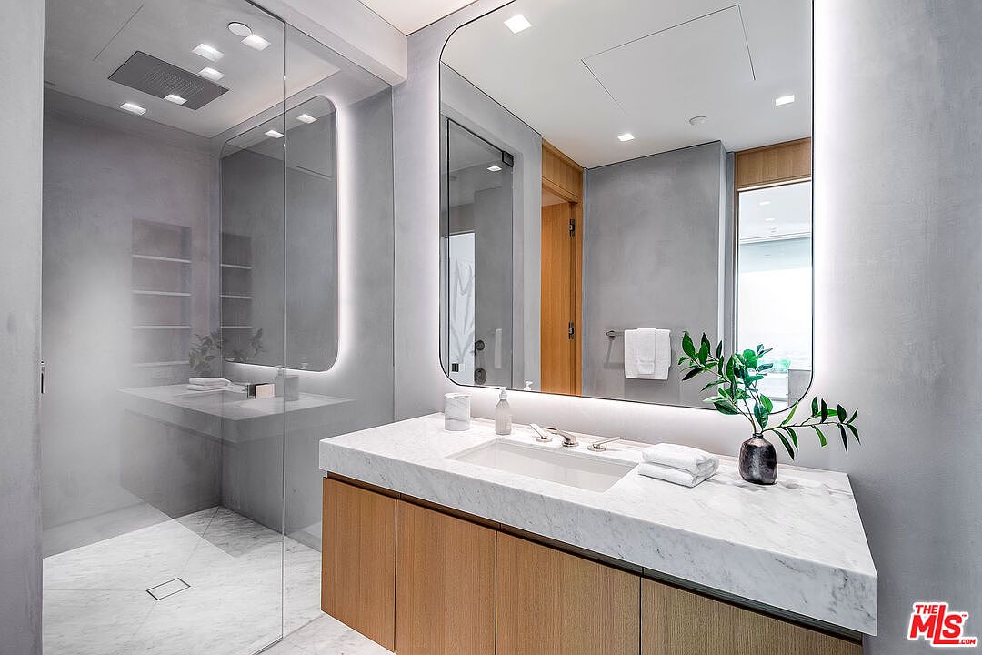 This modern primary bathroom features a sleek vanity with a white marble countertop and warm wood cabinetry, complemented by a large, backlit rounded-corner mirror. To the left, a spacious walk-in shower with a glass enclosure and a rainfall showerhead adds a luxurious, spa-like feel to the room. The overall aesthetic is clean, contemporary, and sophisticated, utilizing a neutral color palette to create a serene atmosphere.